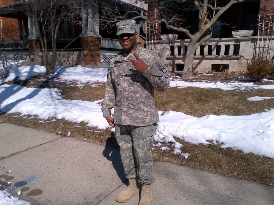 Soldier in camouflage uniform standing on a sidewalk, making a peace sign with fingers, smiling with snow on the ground behind him.