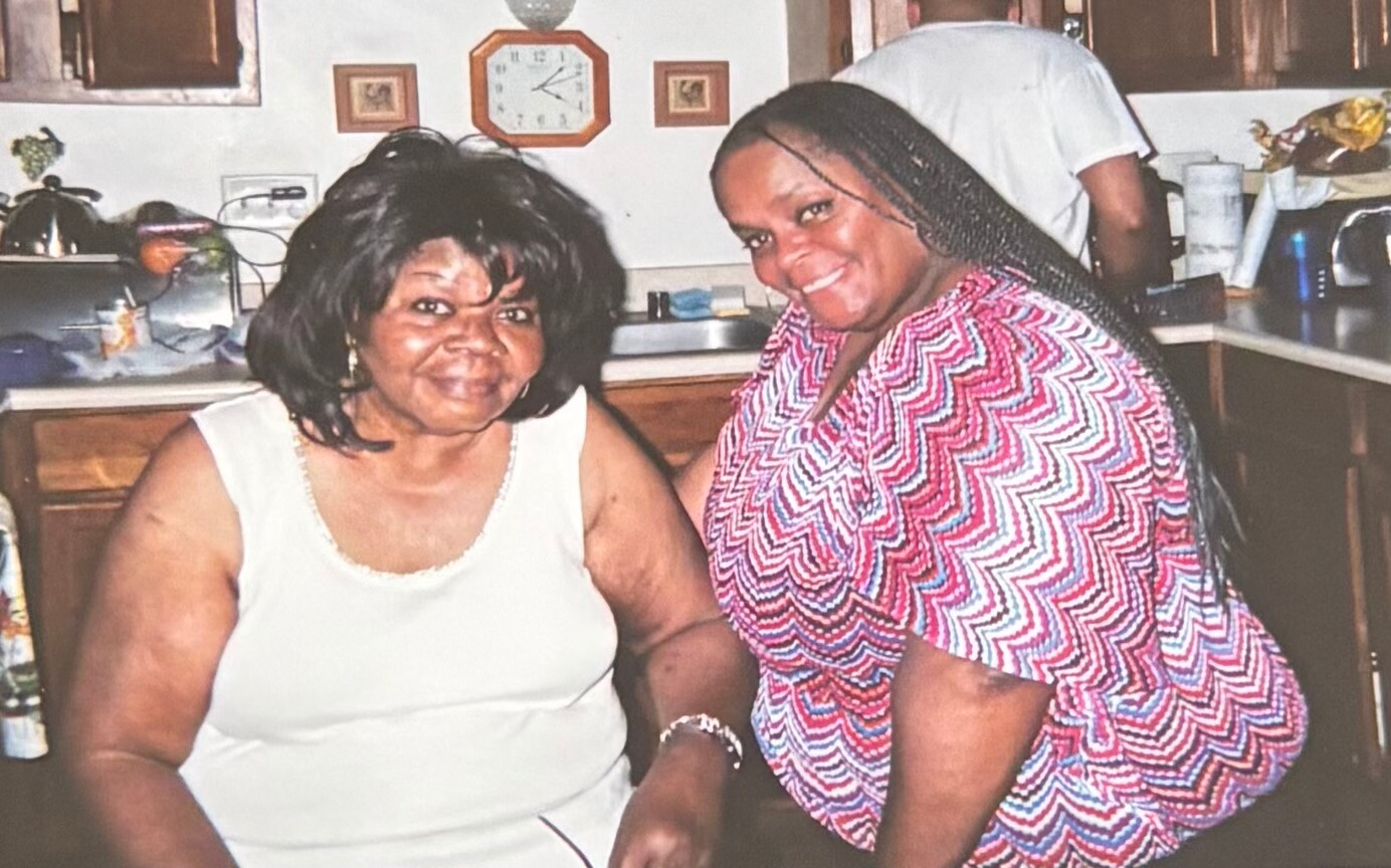 Two women smiling and posing in a home kitchen; one seated in a white sleeveless top, the other in a pink zigzag blouse.