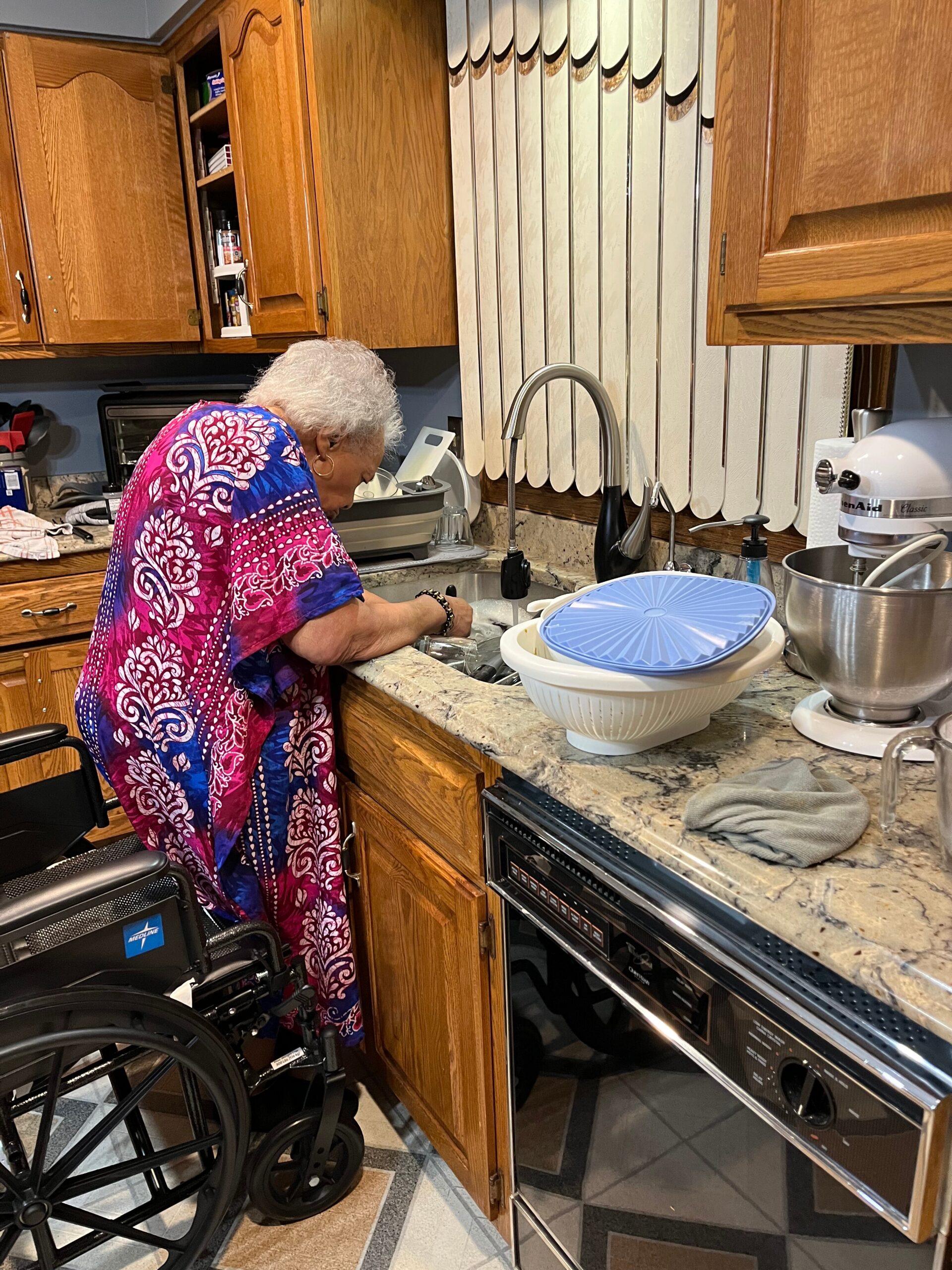 Older woman washing dishes at a kitchen sink, wearing a bright pink and blue patterned garment, with a wheelchair nearby.