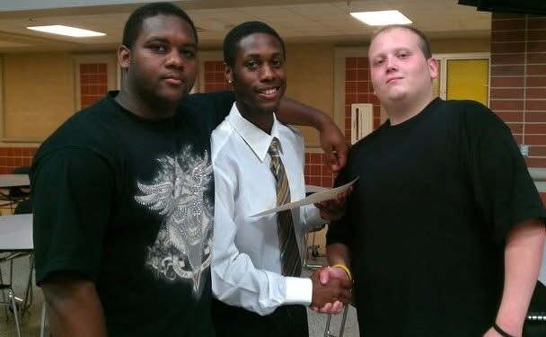 Three male students in a school cafeteria; the middle student in a white shirt and tie shakes hands with the student on the right while the left student stands nearby.