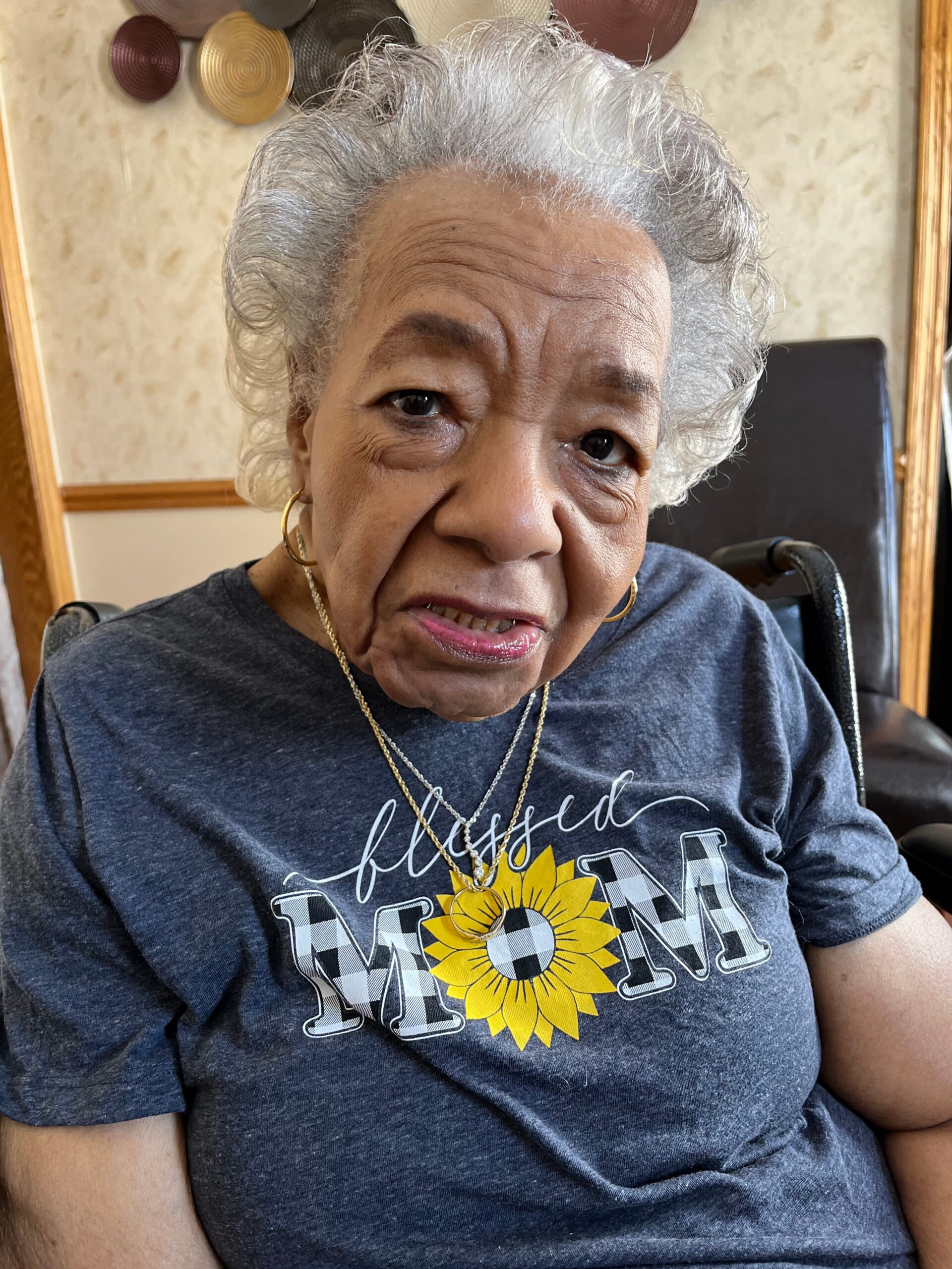 Close-up of smiling elderly African American woman in a wheelchair, wearing a dark T-shirt with a yellow sunflower graphic and bold lettering nearby.