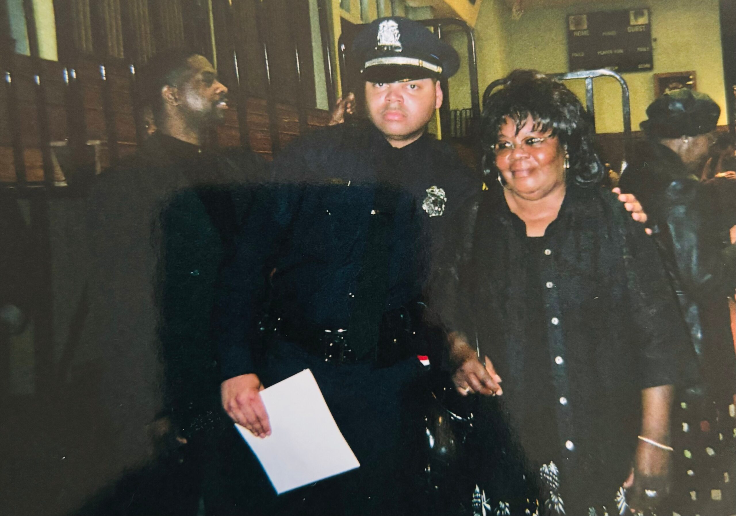 Police officer with hat posing for a photo with a smiling woman indoors, other people in the background.