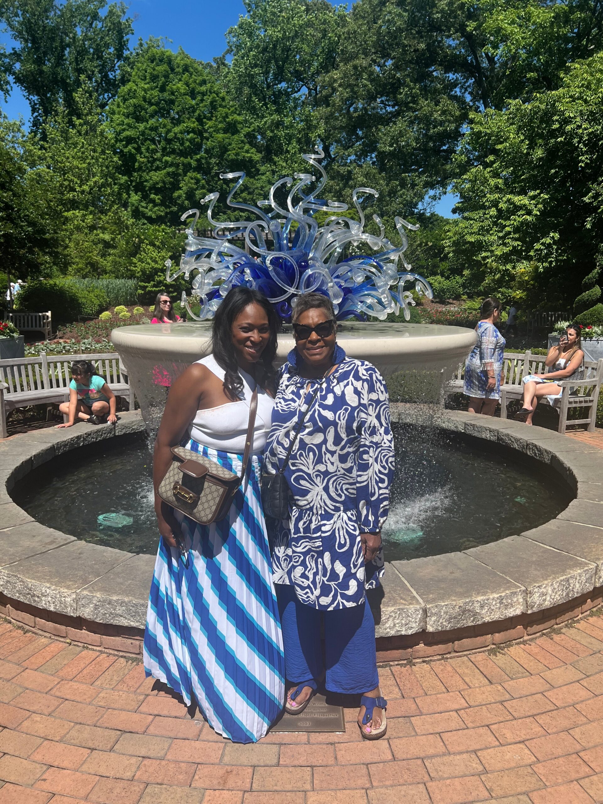 Two women pose together in front of a blue glass sculpture fountain in a sunny garden courtyard.