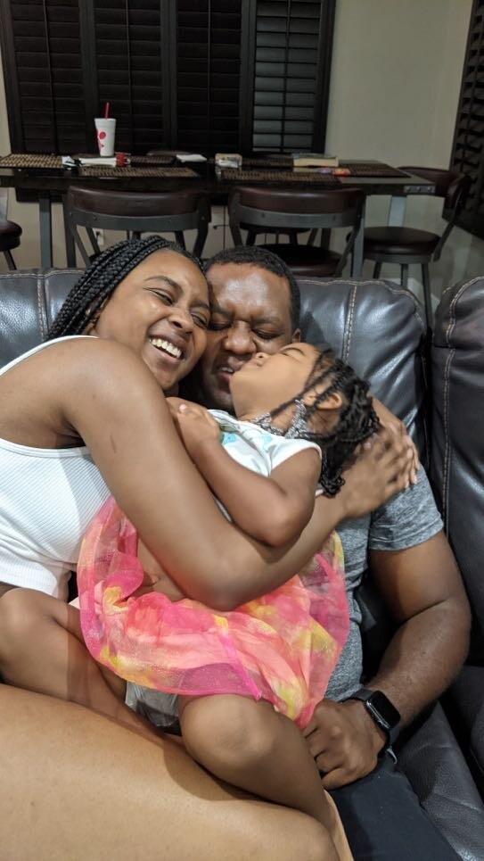 A joyful Black family cuddles on a dark leather couch: mother in a white top, father beside her, cradling their young daughter in a pink dress.