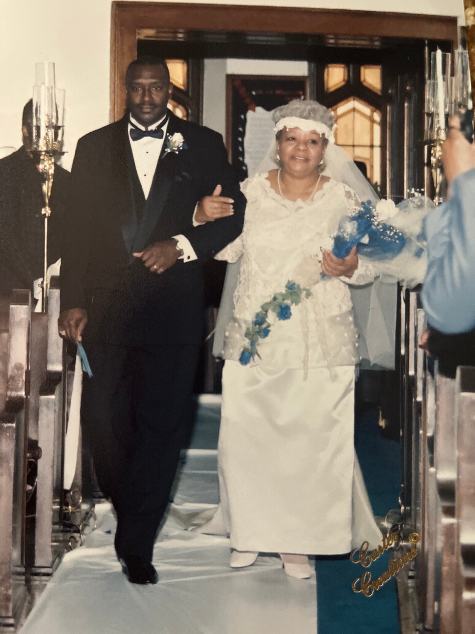 Bride and groom walking down church aisle after ceremony, she in white gown with veil and blue bouquet, he in black tuxedo.