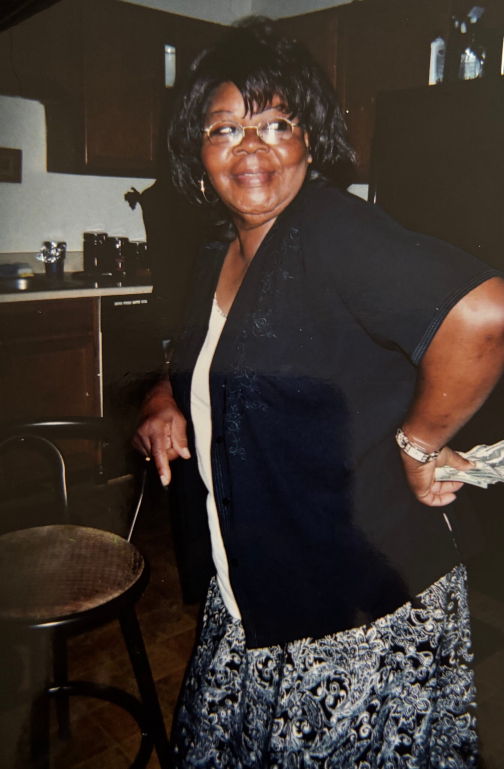 Smiling woman with glasses and a dark cardigan stands in a kitchen, holding cash behind her back.