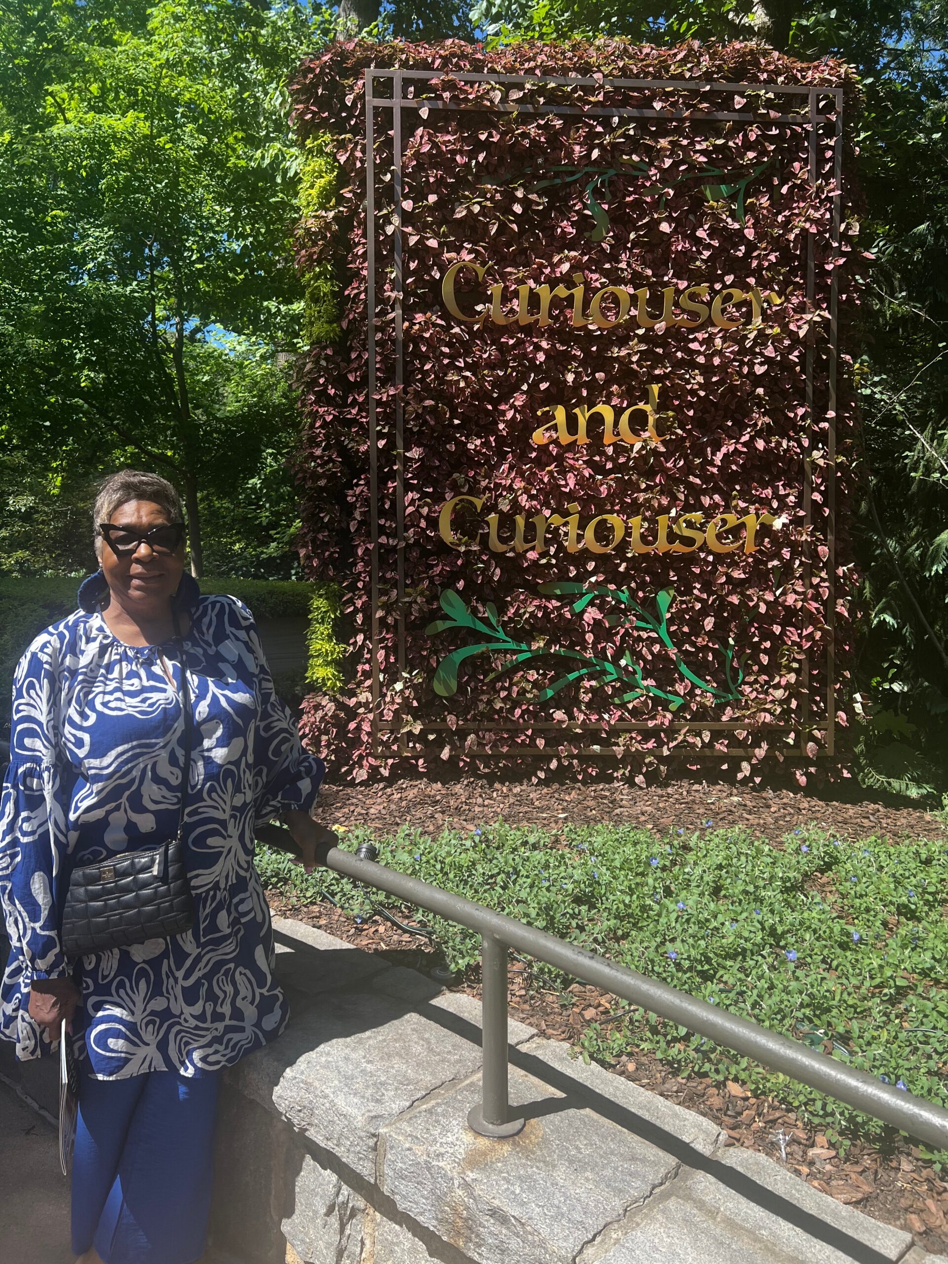 Woman in a blue patterned dress and sunglasses stands beside a decorative sign reading 'Curious and Curiouser' covered in burgundy leaves.