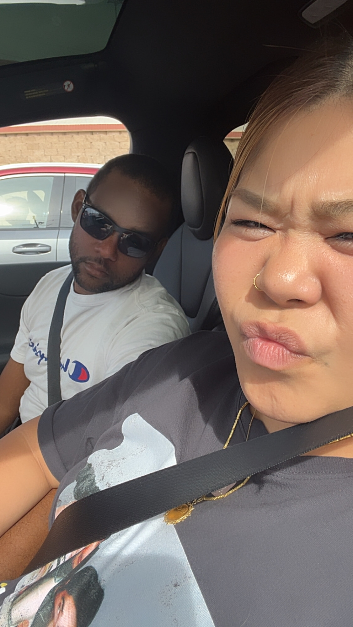 Close-up of a woman wearing a nose ring in a car, squinting at the camera with a seatbelt on, while a man in sunglasses sits behind her.