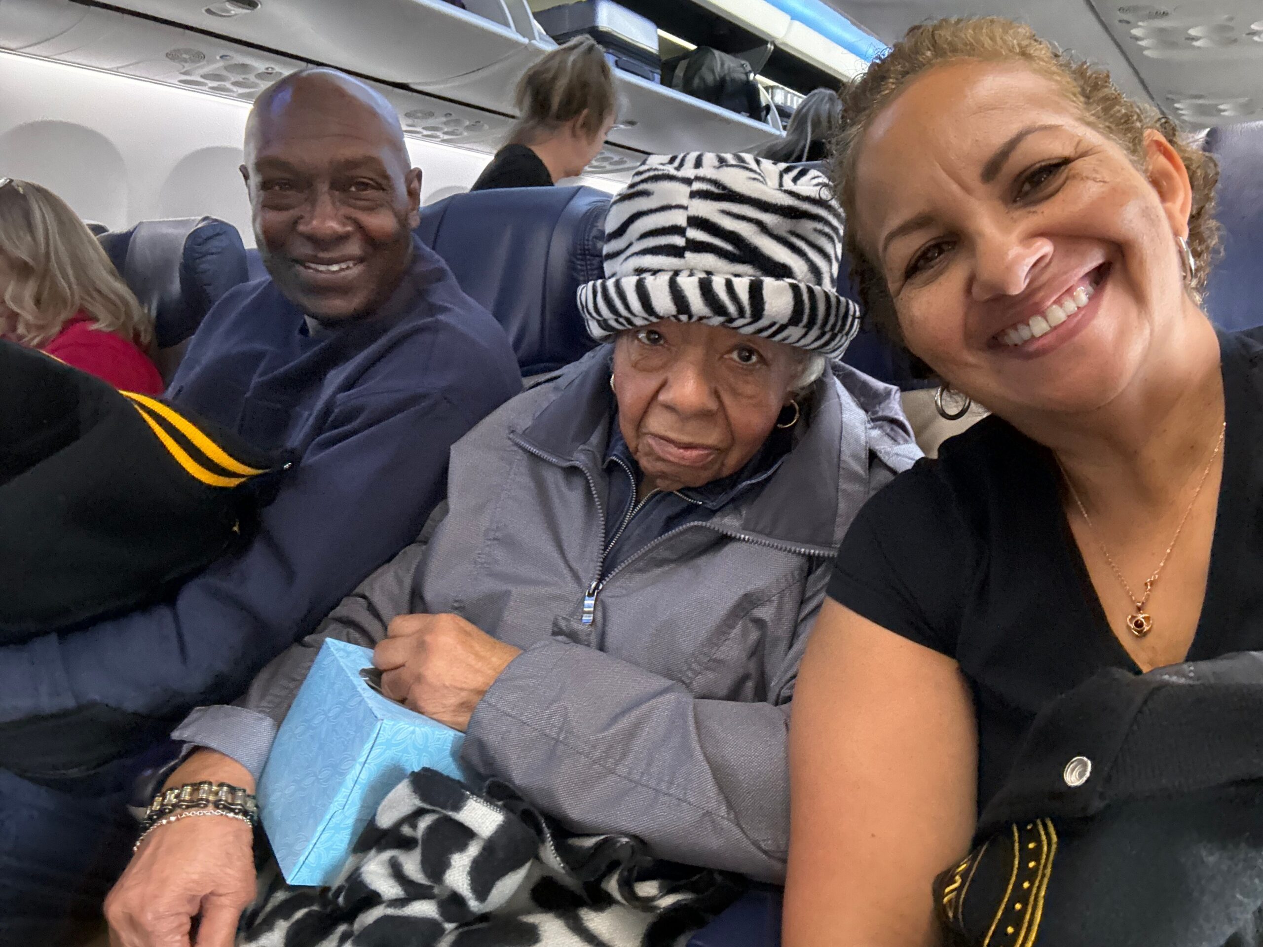 Smiling group of passengers on a airplane, taking a close-up selfie in a cabin with overhead bins visible in the background.