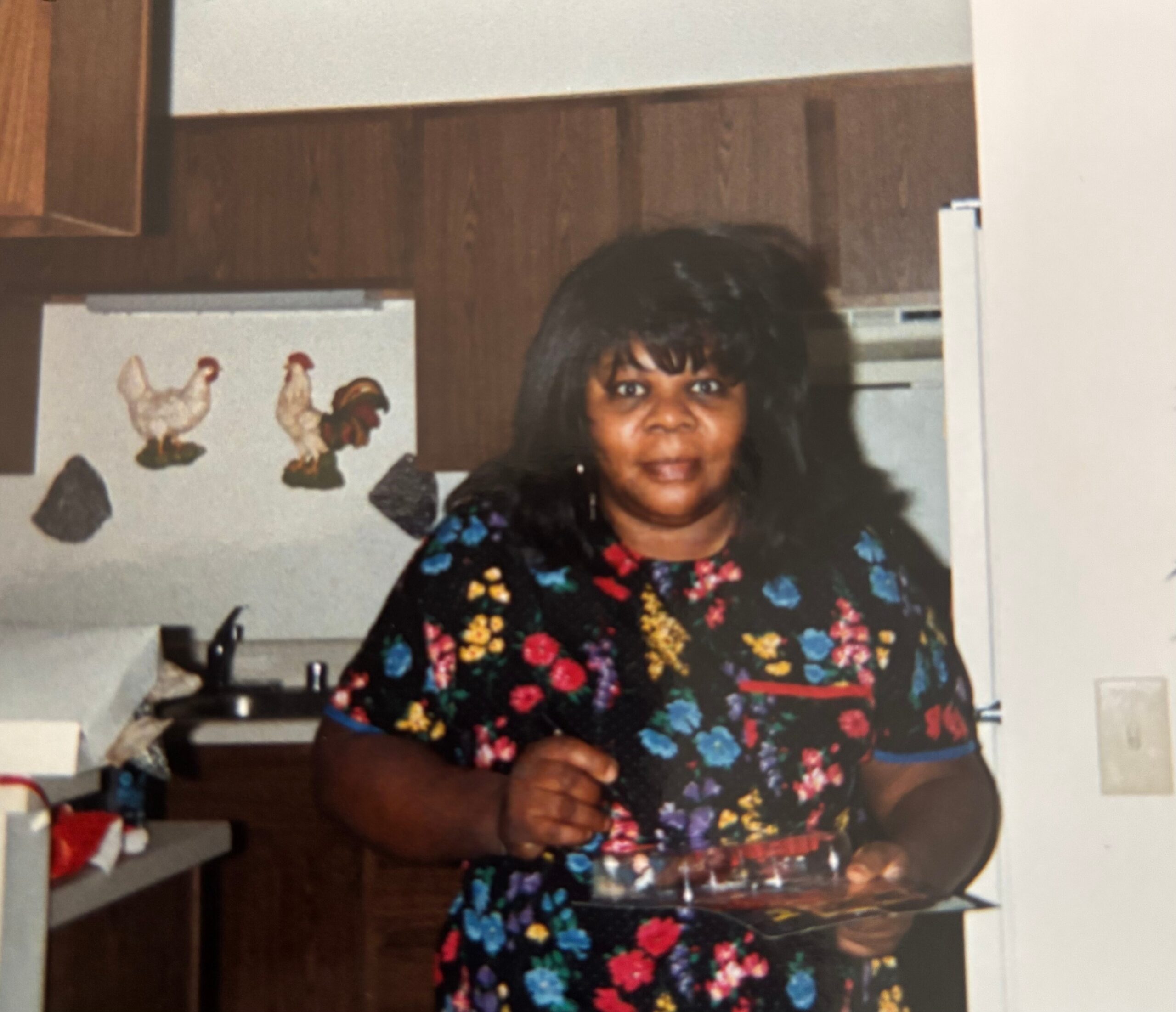 Smiling woman with dark hair wearing a colorful floral dress indoors, near a kitchen workspace.