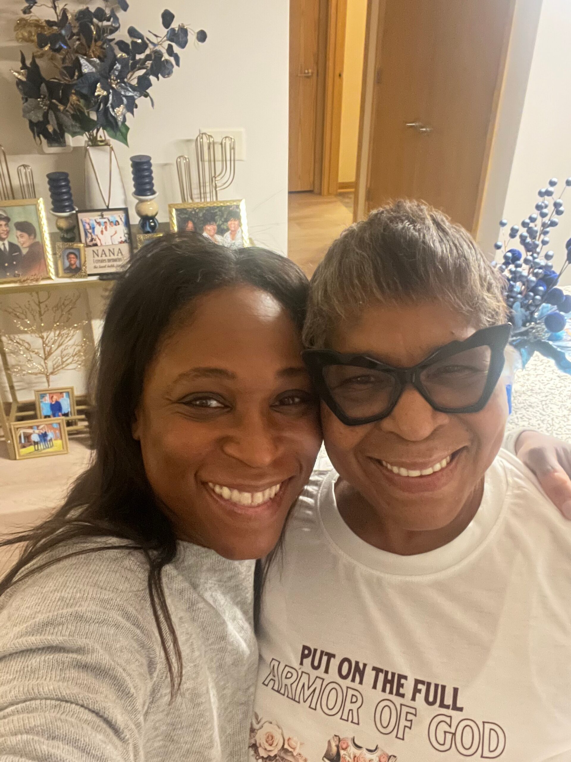 Two women take a cheerful selfie indoors, faces close together and smiling; background shows a shelf with family photos and decorative items.