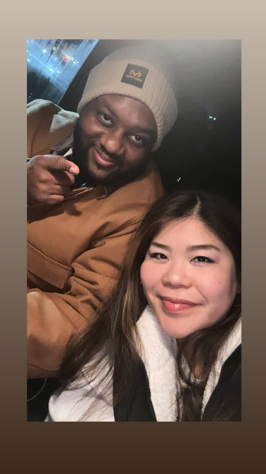Smiling couple taking a selfie in a car at night; man in a beige beanie points at the camera, woman in the foreground wears a scarf.