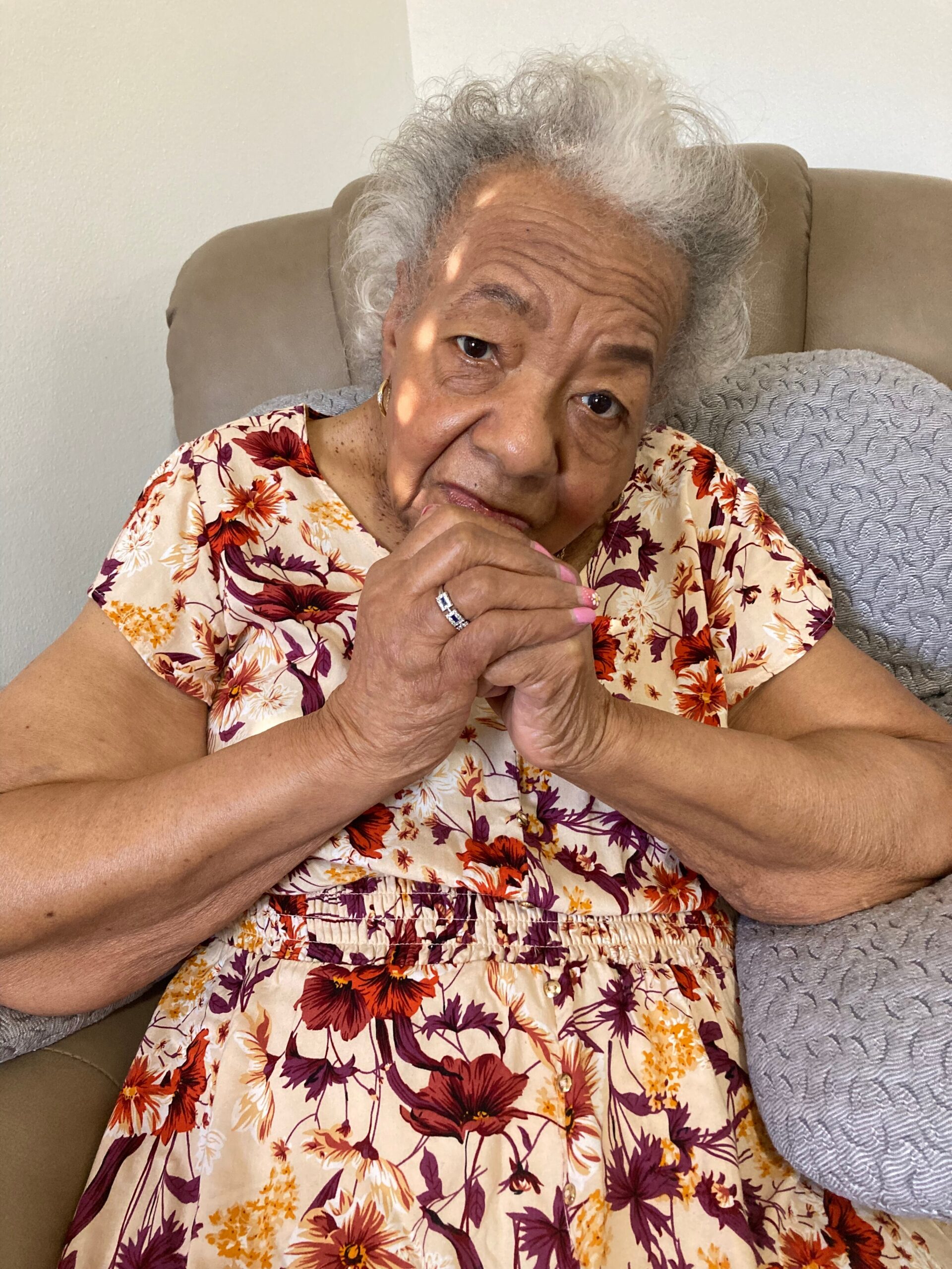 Older woman with gray hair sits on a beige couch, hands clasped in her lap, wearing a cream dress with orange, purple, and yellow floral pattern.