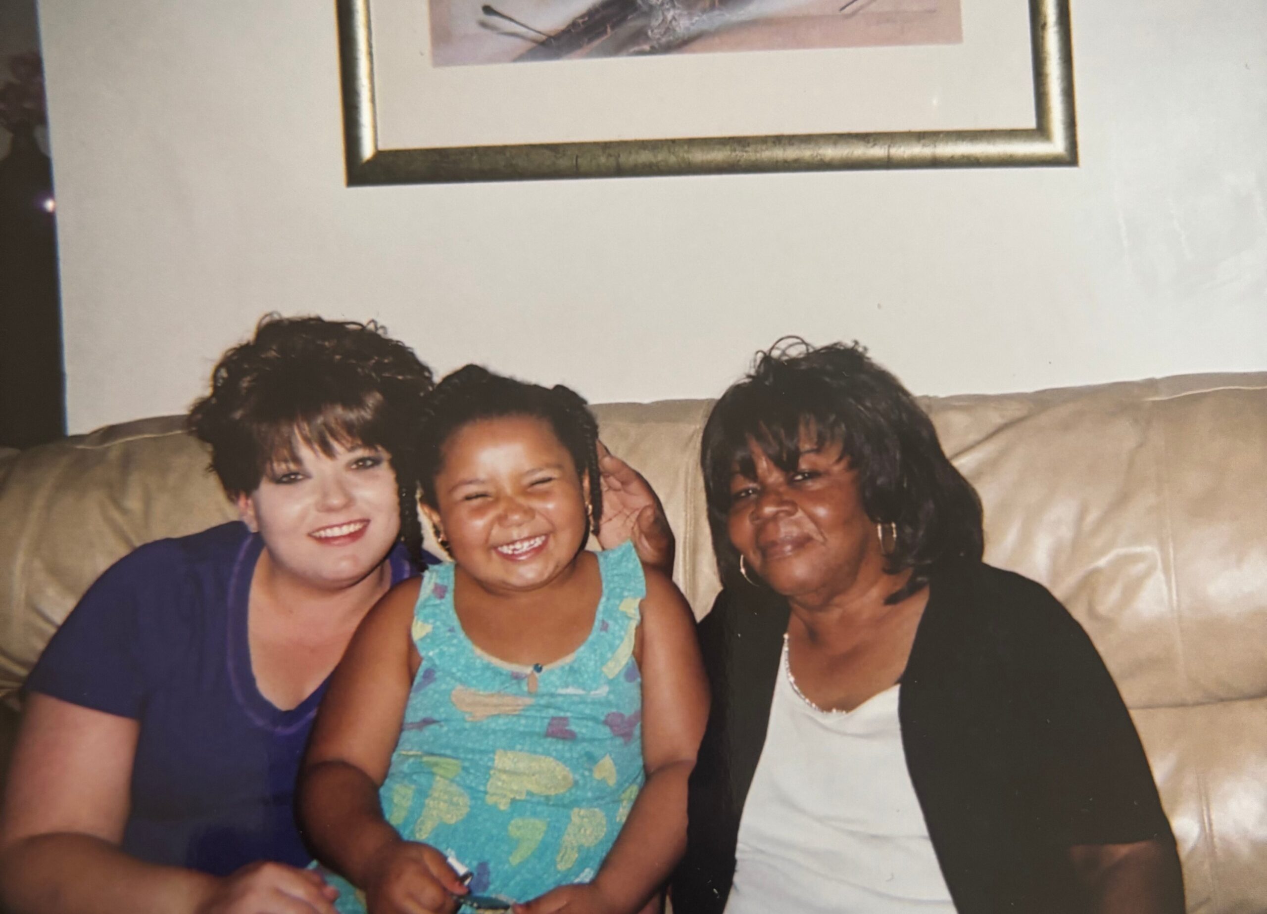 Three women sit together on a beige couch, smiling for the photo with arms around each other.