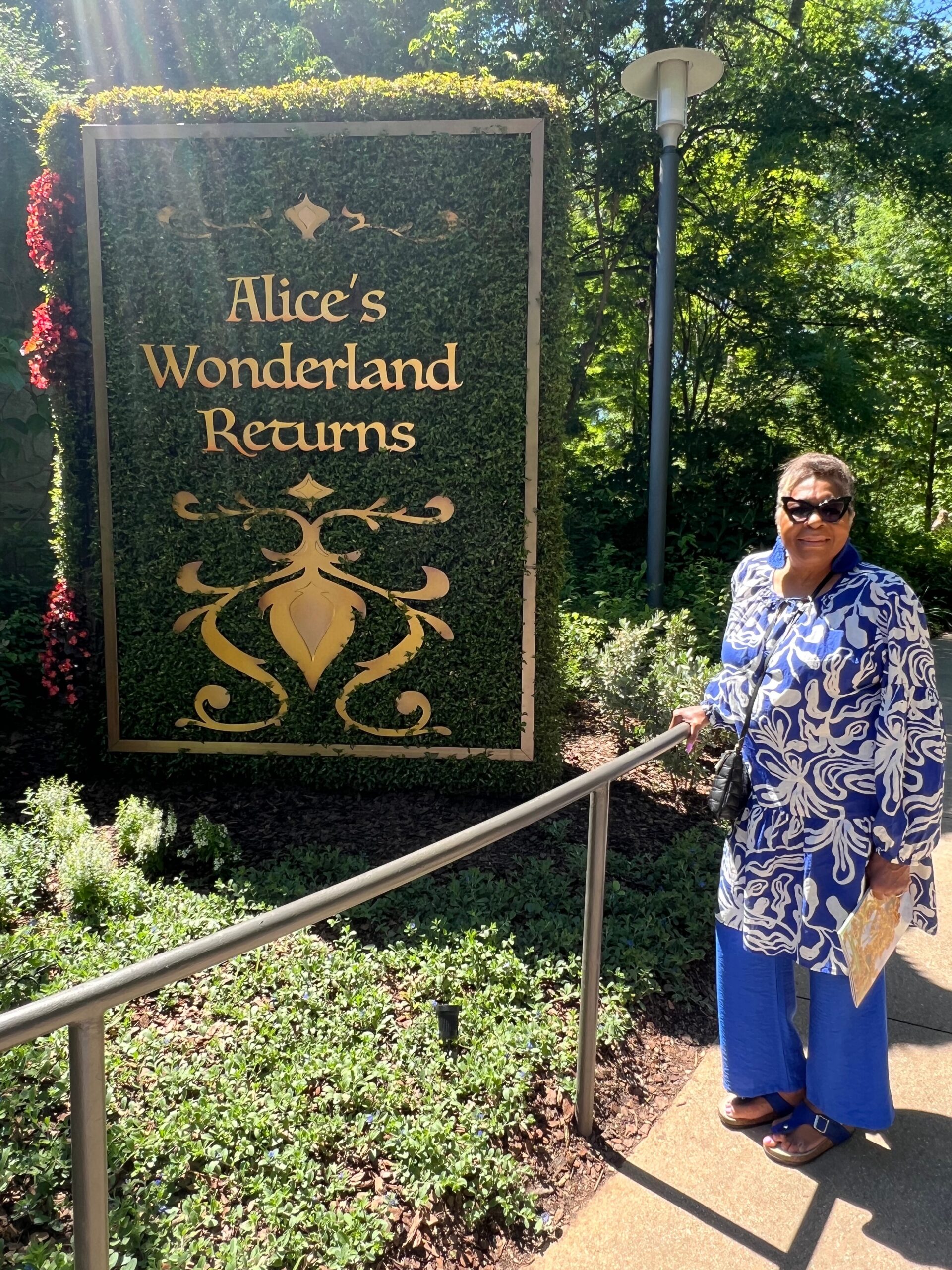 Sign reading 'Alice's Wonderland Returns' in gold letters against green hedging, decorative gold motif beneath, outdoors with sunlit foliage.