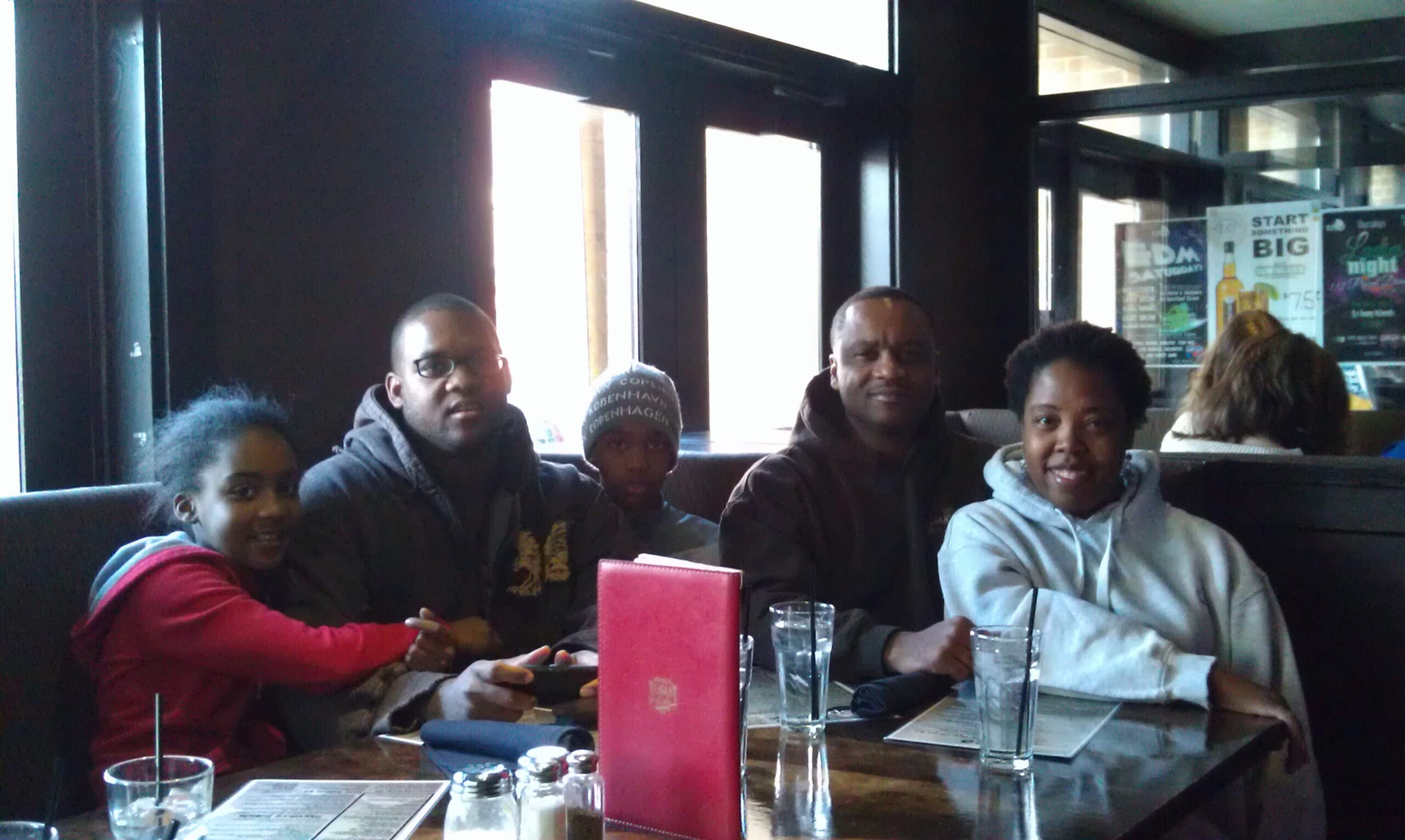 Group of five people seated in a restaurant booth, smiling at the camera with drinks on the table in front of them.
