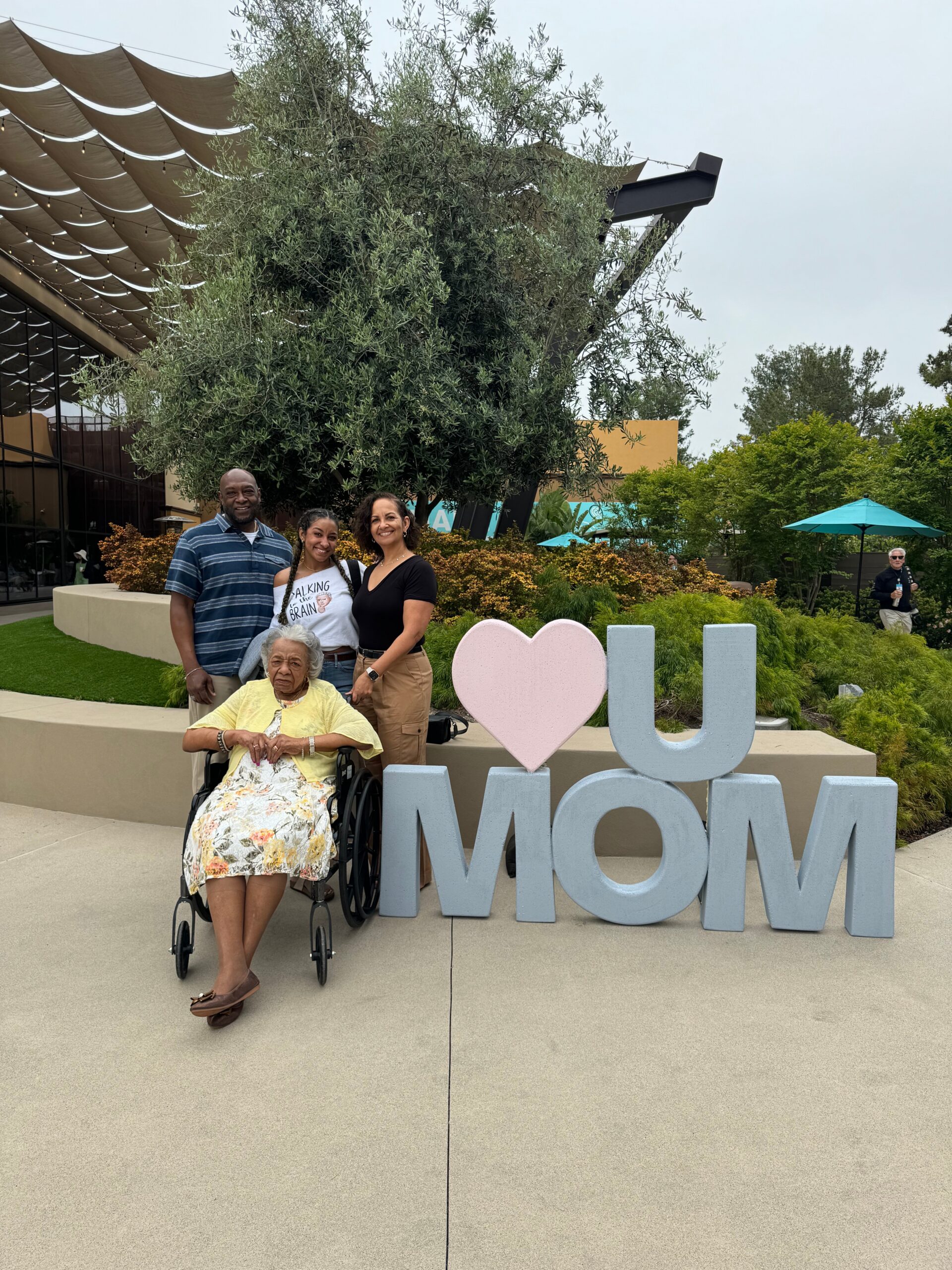 Family posing outdoors beside a large 'I heart Mom' sculpture, with an elderly woman in a wheelchair at the front.