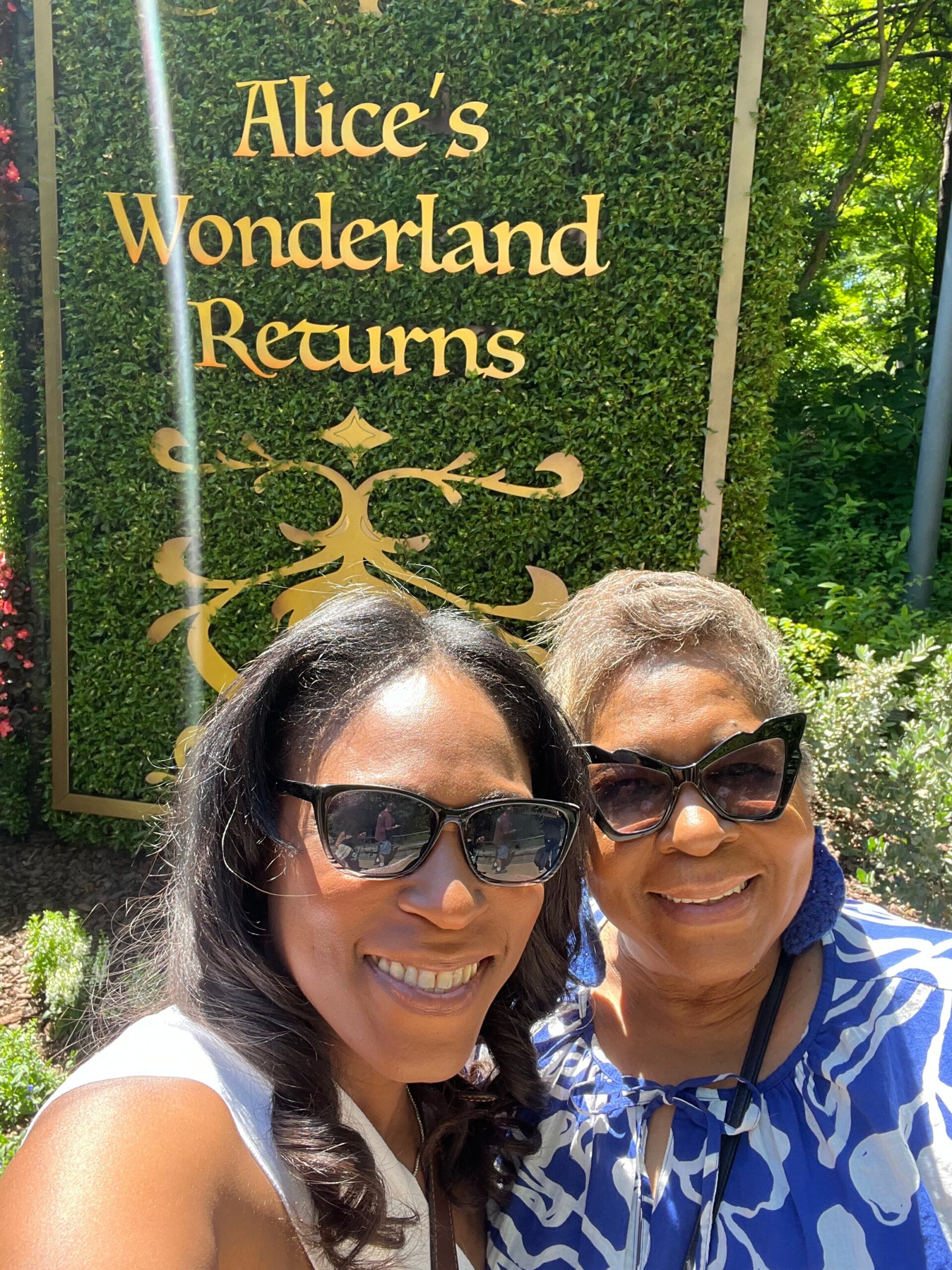 Two women smiling for a selfie in front of a decorative hedge sign reading 'Alice's Wonderland Returns' in gold letters.