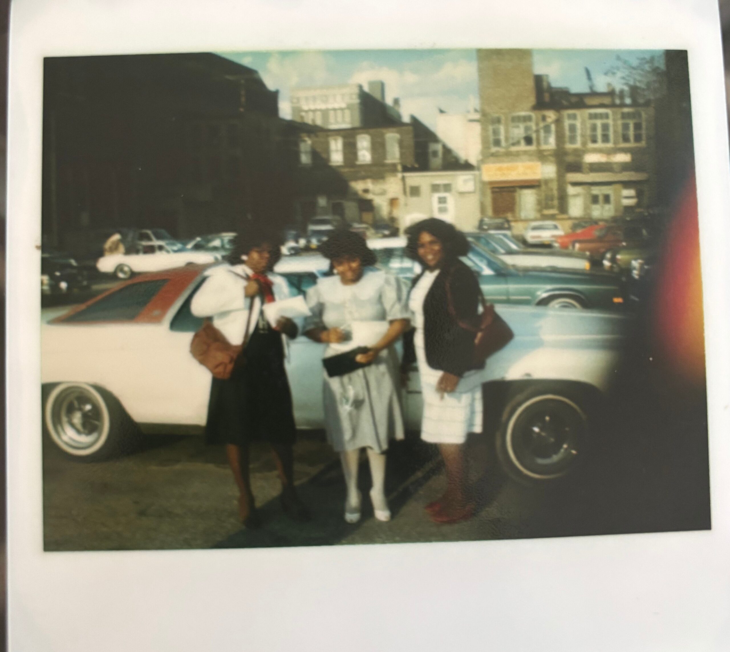 Three young women posing together in front of vintage cars on a city street, with brick buildings in the background.