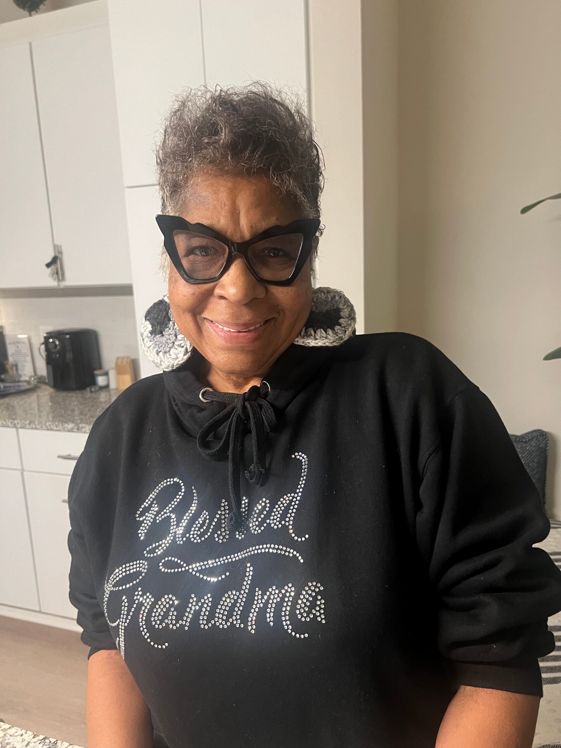 Smiling woman with short gray hair wearing oversized black cat-eye sunglasses and a rhinestone-printed black hoodie in a kitchen setting.