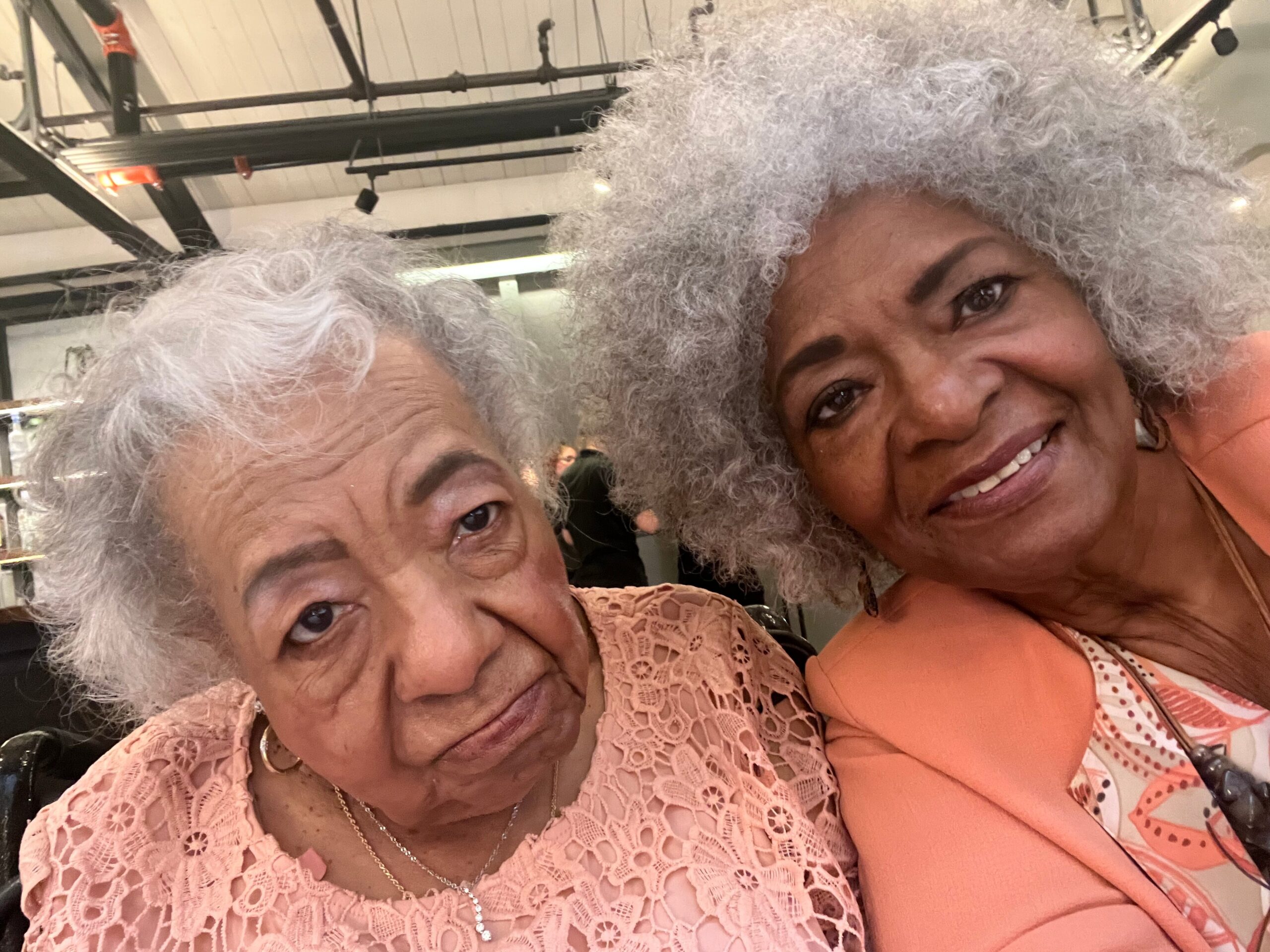 Two elderly women with gray hair posing for a close-up selfie in a store, both smiling softly, wearing peach lace tops.