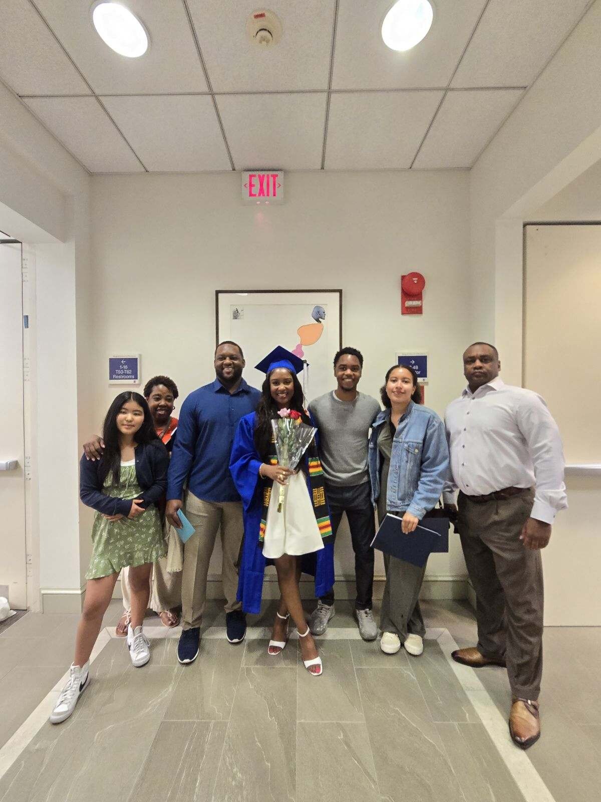 Group of seven people posing in a hallway; one graduate in blue cap and gown holding flowers, flanked by friends and family.