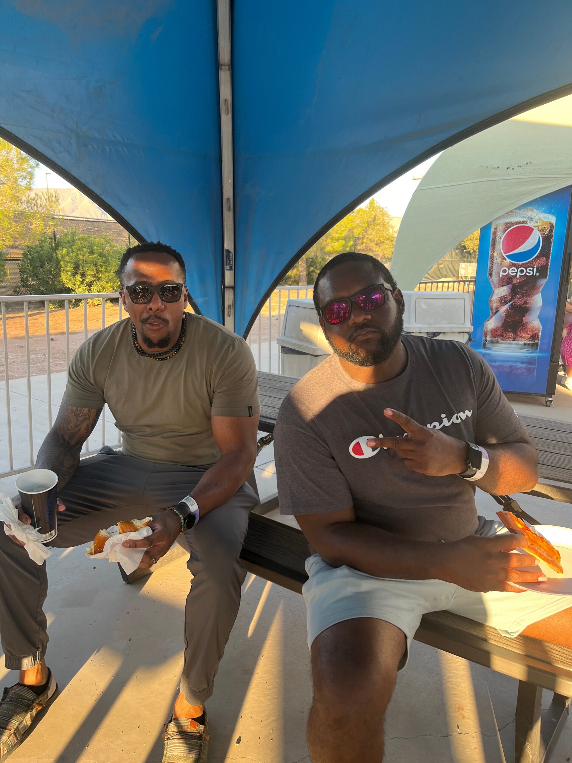 Two men wearing sunglasses sit at a picnic table under a blue canopy, eating hot dogs with a Pepsi banner visible in the background.