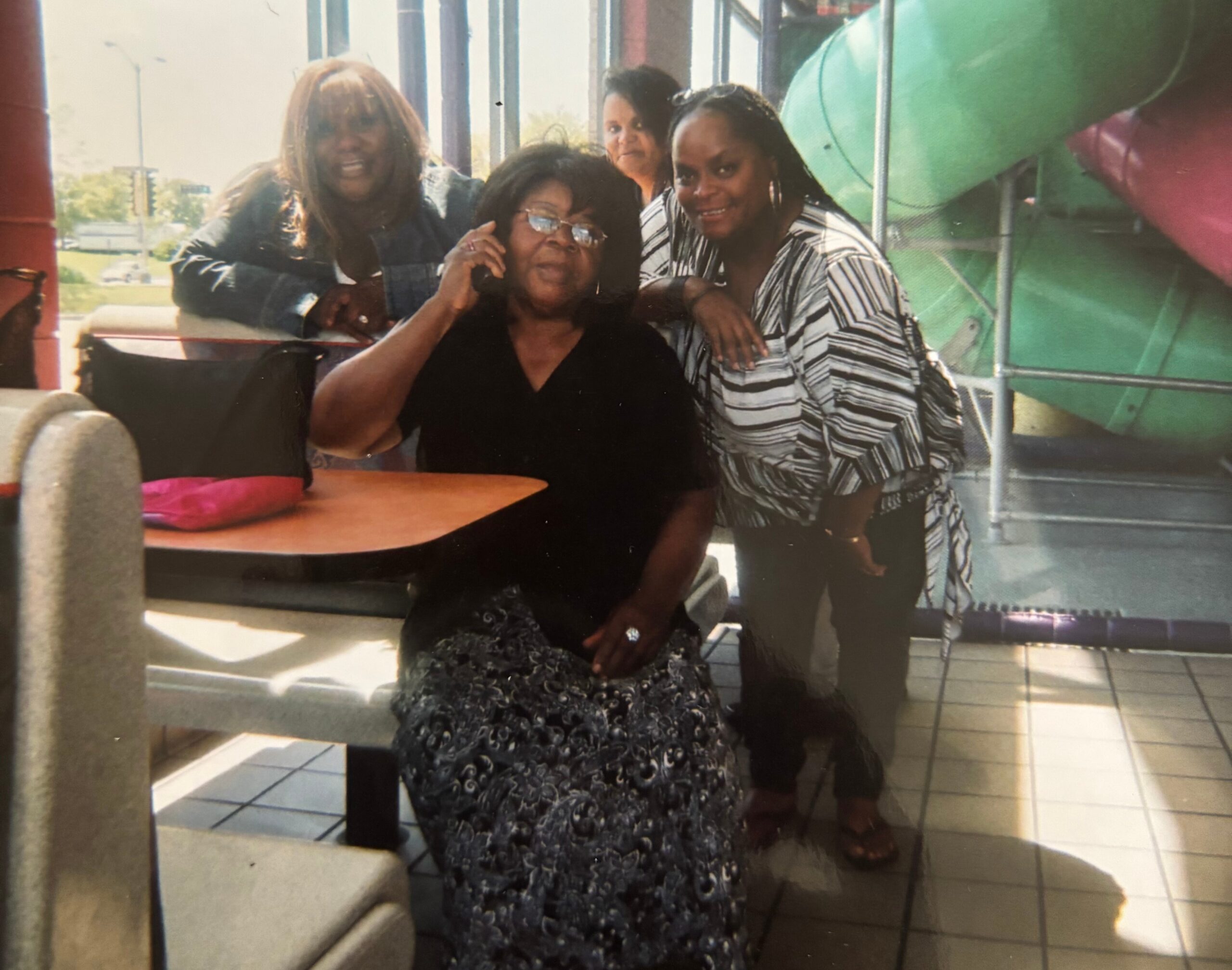 Group of four women posing together indoors, one on a phone, smiling at the camera near a table and playground equipment in the background