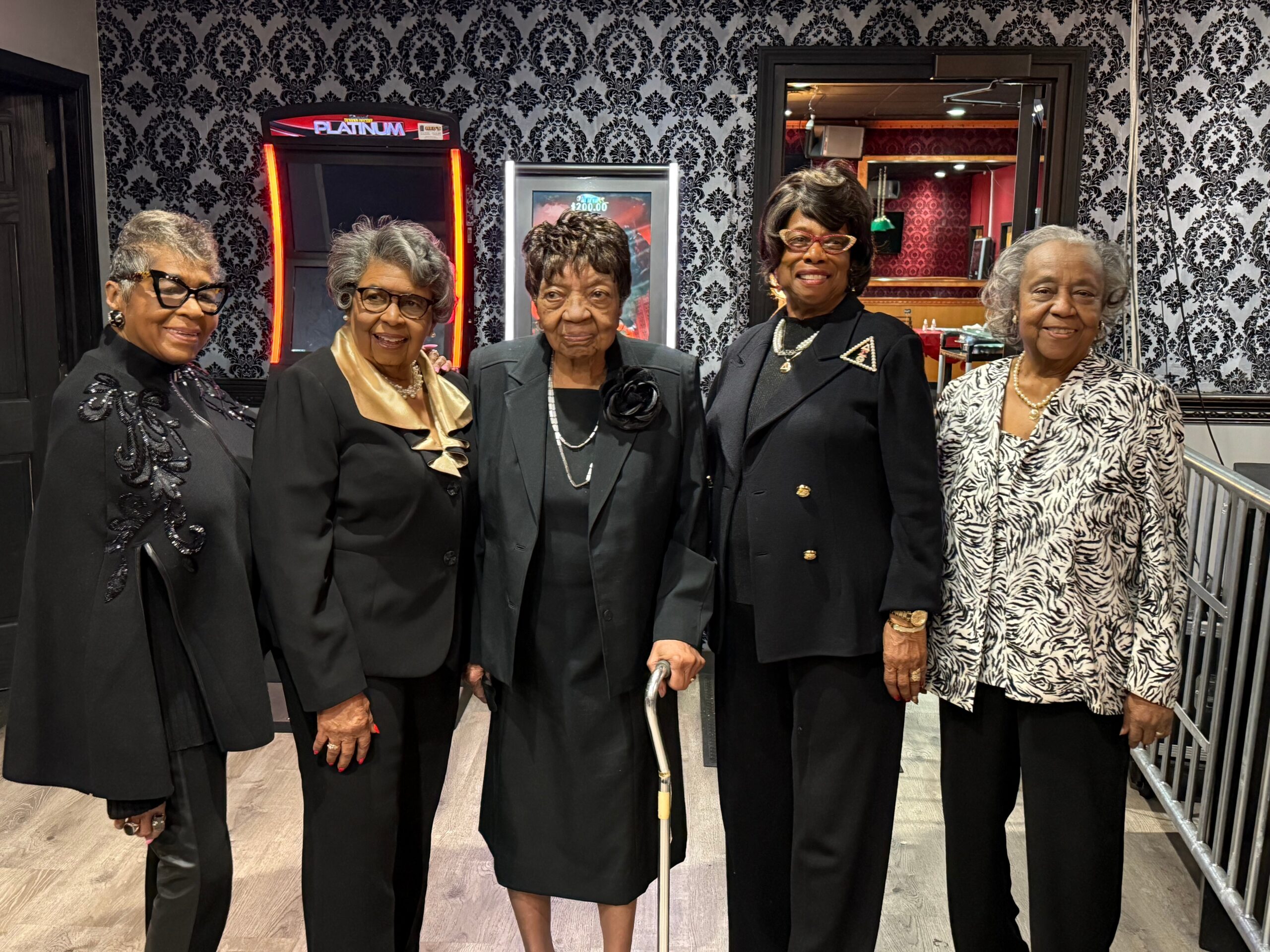 Group of five elderly women in formal black outfits posing together in a room with damask wallpaper and a doorway in the background.