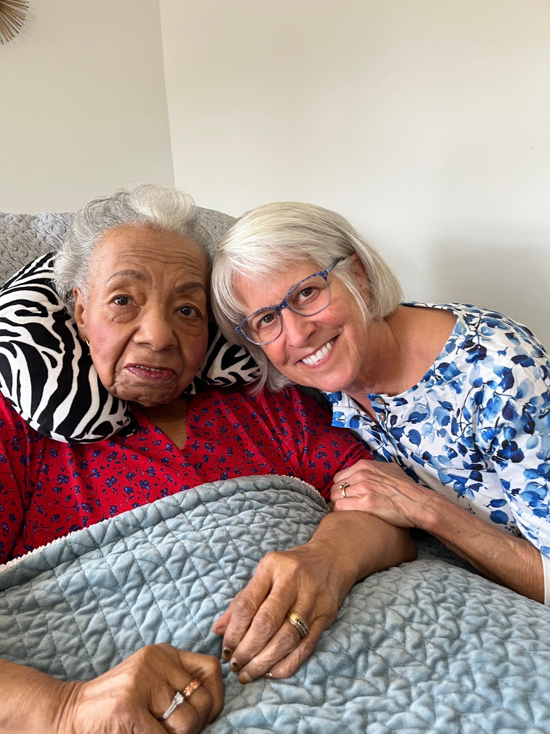 Two elderly women sit closely on a couch, smiling, with their hands interlaced over a light blue quilt. The woman on the left wears a red floral blouse, the woman on the right wears a blue floral shirt and glasses.