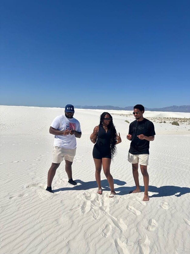 Three friends pose on white desert sand under a clear blue sky, with distant mountains on the horizon.