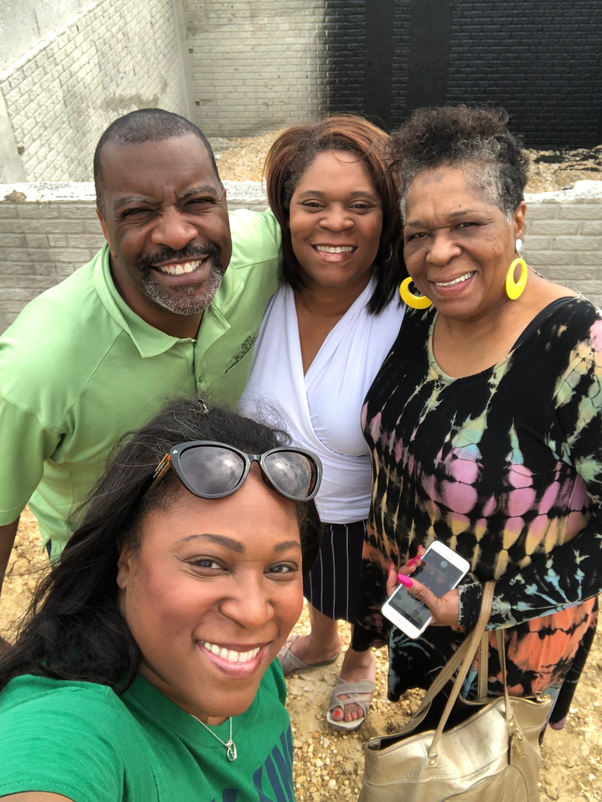 Four adults pose for a cheerful outdoor selfie, smiling at the camera with brick walls in the background.