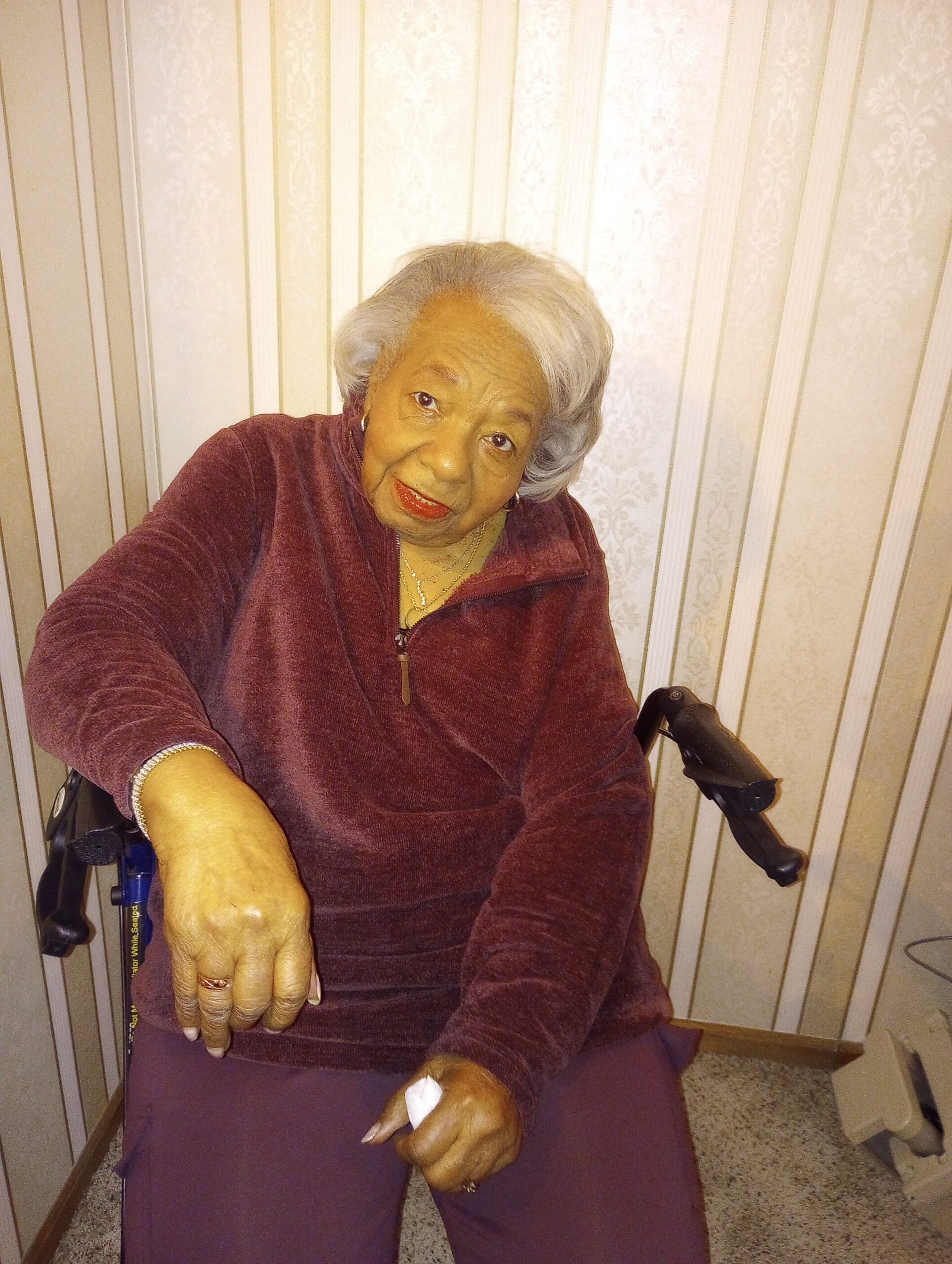 Elderly woman with short gray hair in a burgundy top, seated near a walker in a striped beige room.