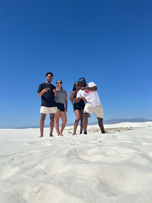 Four friends posing for a selfie on a bright white sandy beach under a clear blue sky, wearing casual summer clothing and sunglasses.