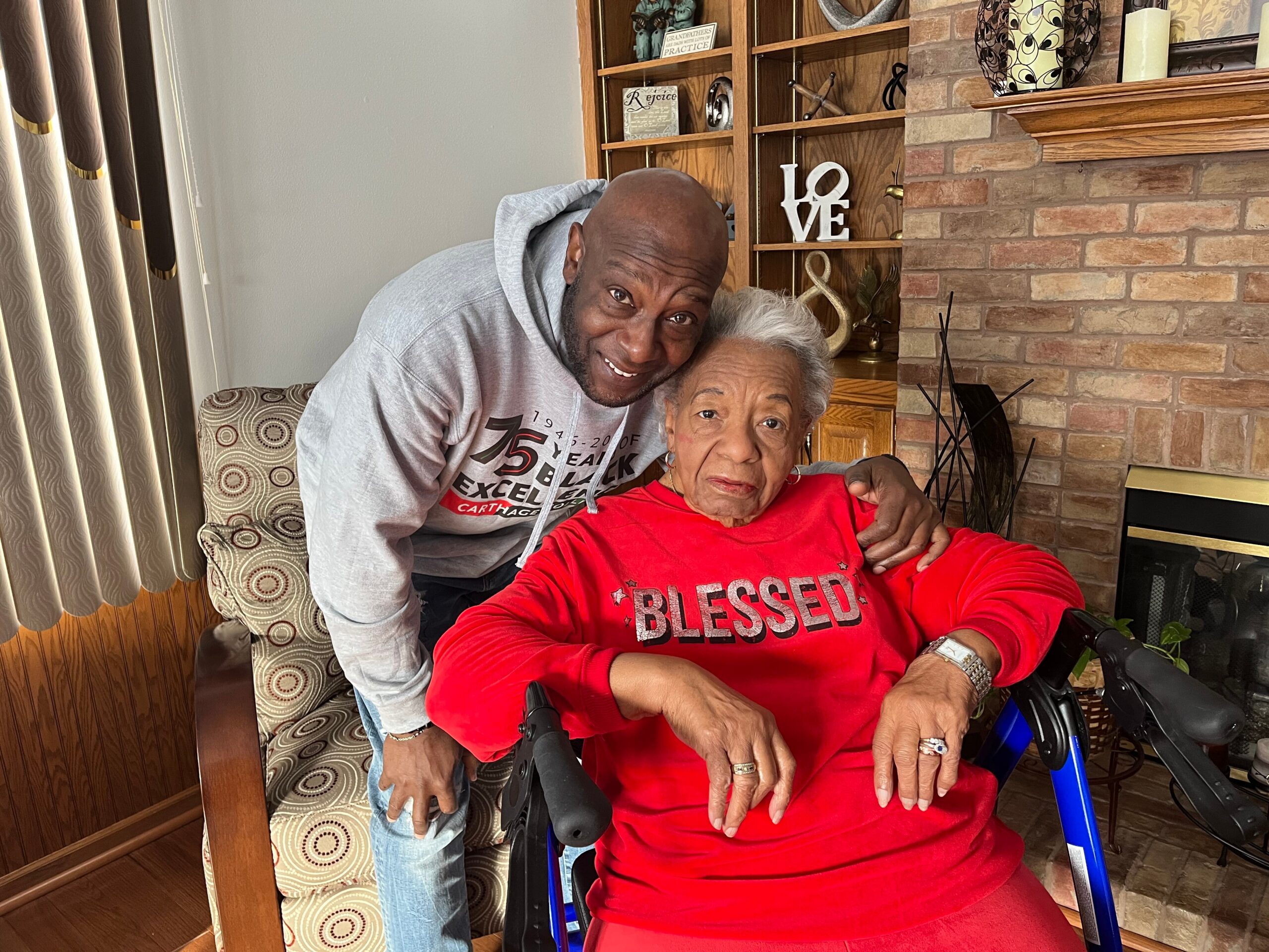 Man leans over and smiles with an elderly woman in a red sweater, seated in a wheelchair indoors beside a brick fireplace.