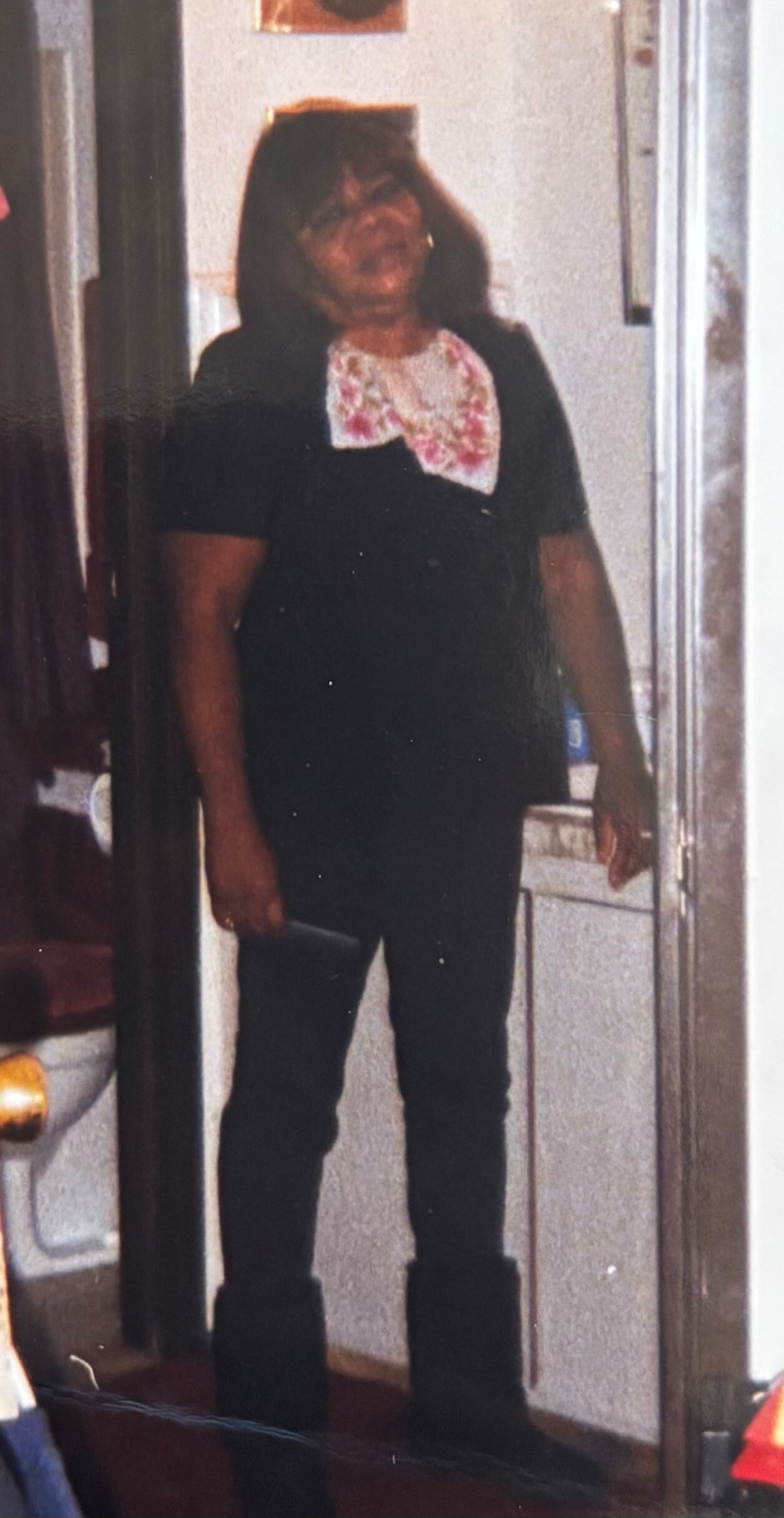 Woman standing in a doorway wearing a black outfit with a pink floral scarf, looking at the camera.