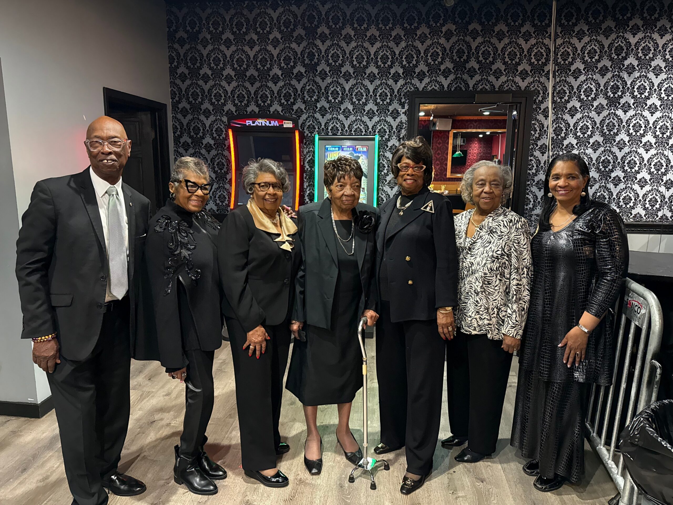 Group of seven adults in formal black attire posing indoors, with arcade games in the background.