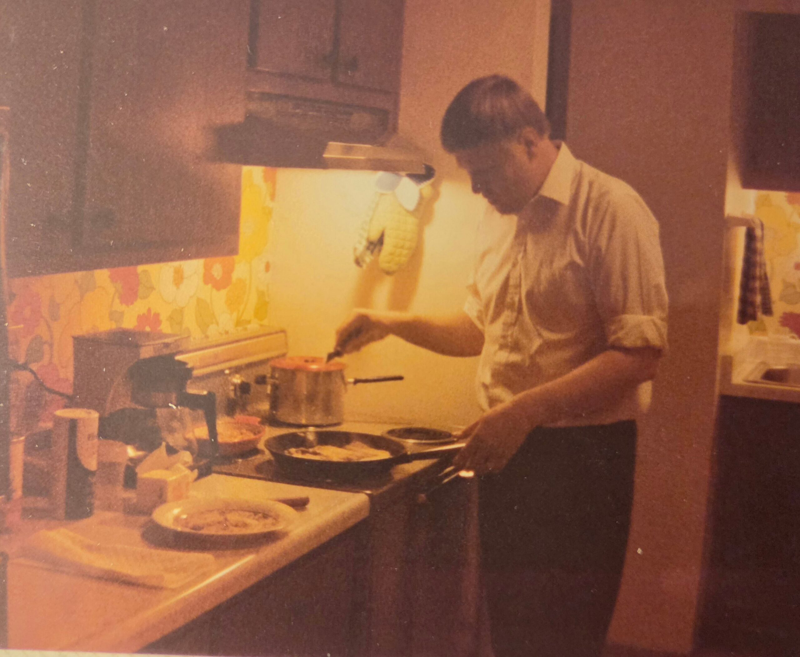 Person stands at a stovetop, stirring a pot in a retro-style kitchen with floral wallpaper in the background.