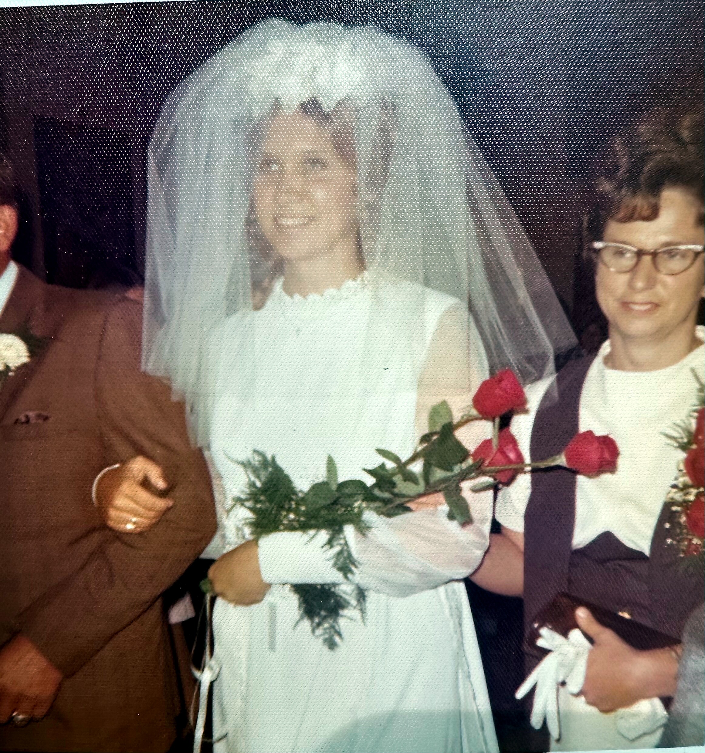 Bride in a white wedding dress and veil holding a bouquet of red roses, standing with a bridesmaid wearing glasses in a party setting.