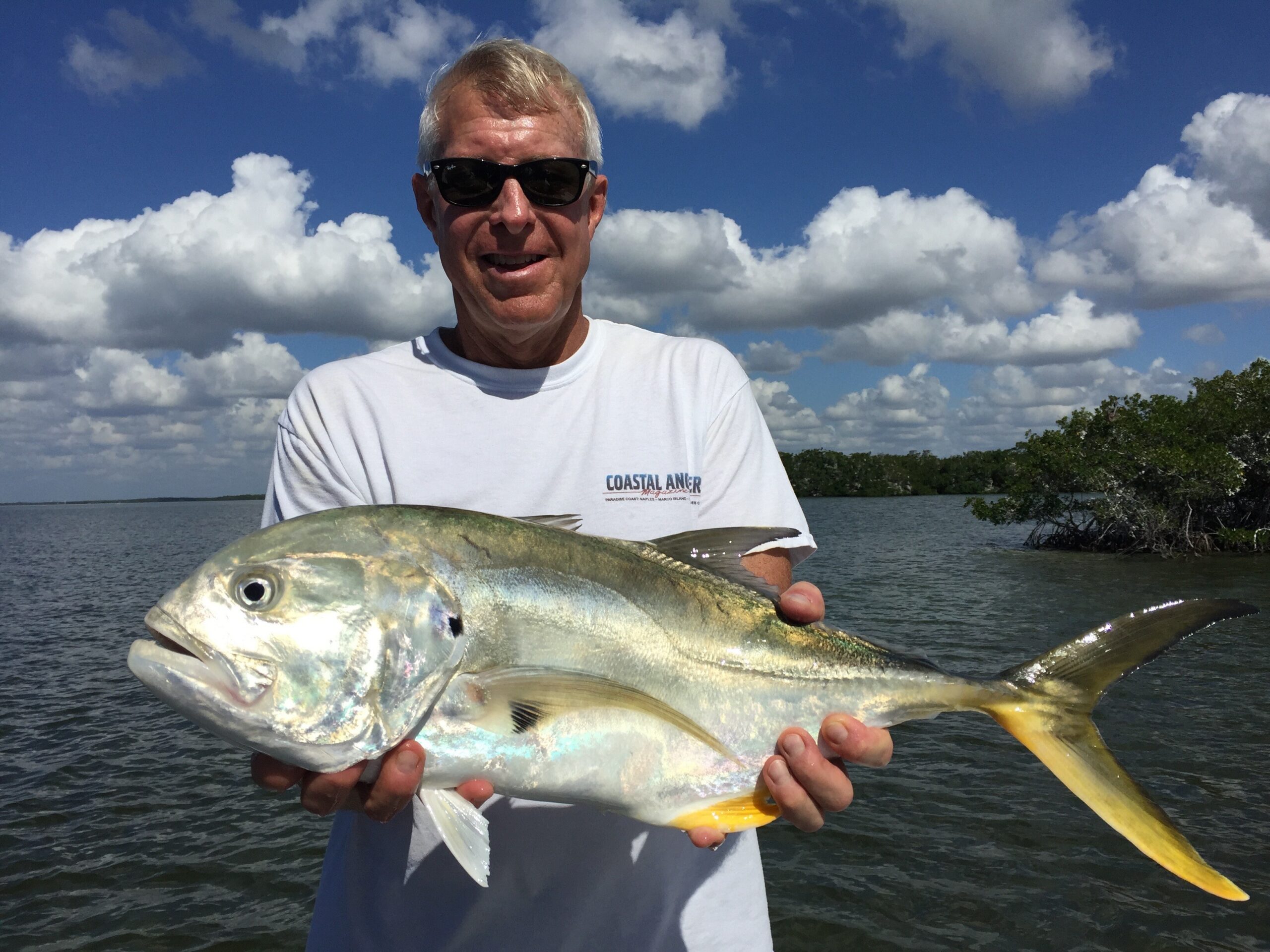Man wearing sunglasses and a white shirt poses with a large yellowtail fish over a body of water on a sunny day.