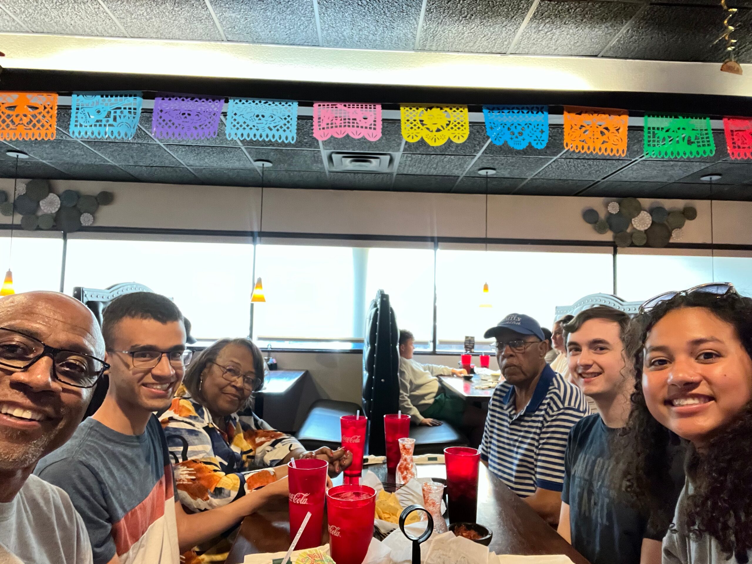 Group of friends at a restaurant taking a selfie, colorful cut-paper banners hanging from the ceiling above them.