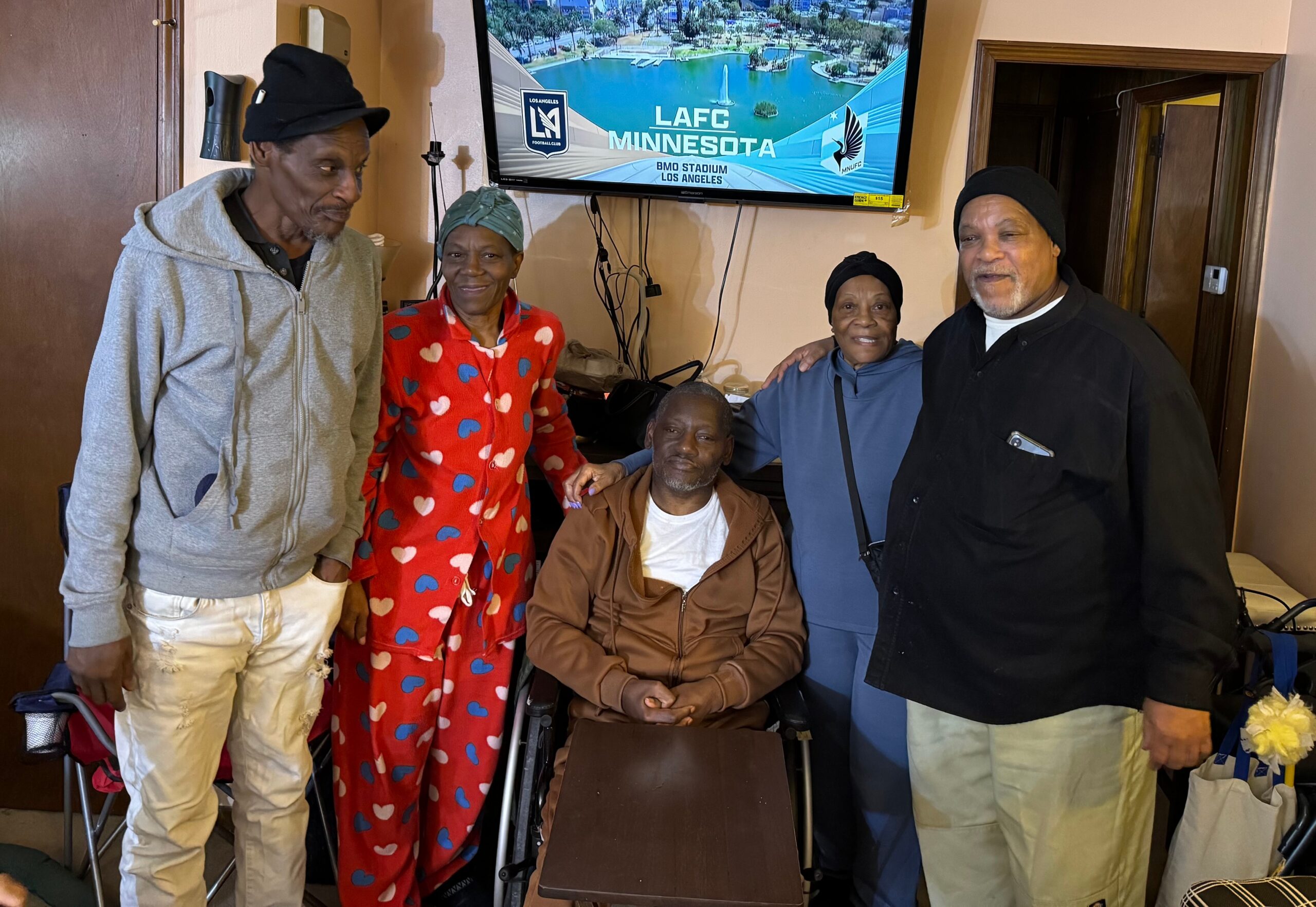 Group of smiling seniors indoors posing for a photo in a living room, with a flat-screen TV displaying a sports graphic on the wall behind them.