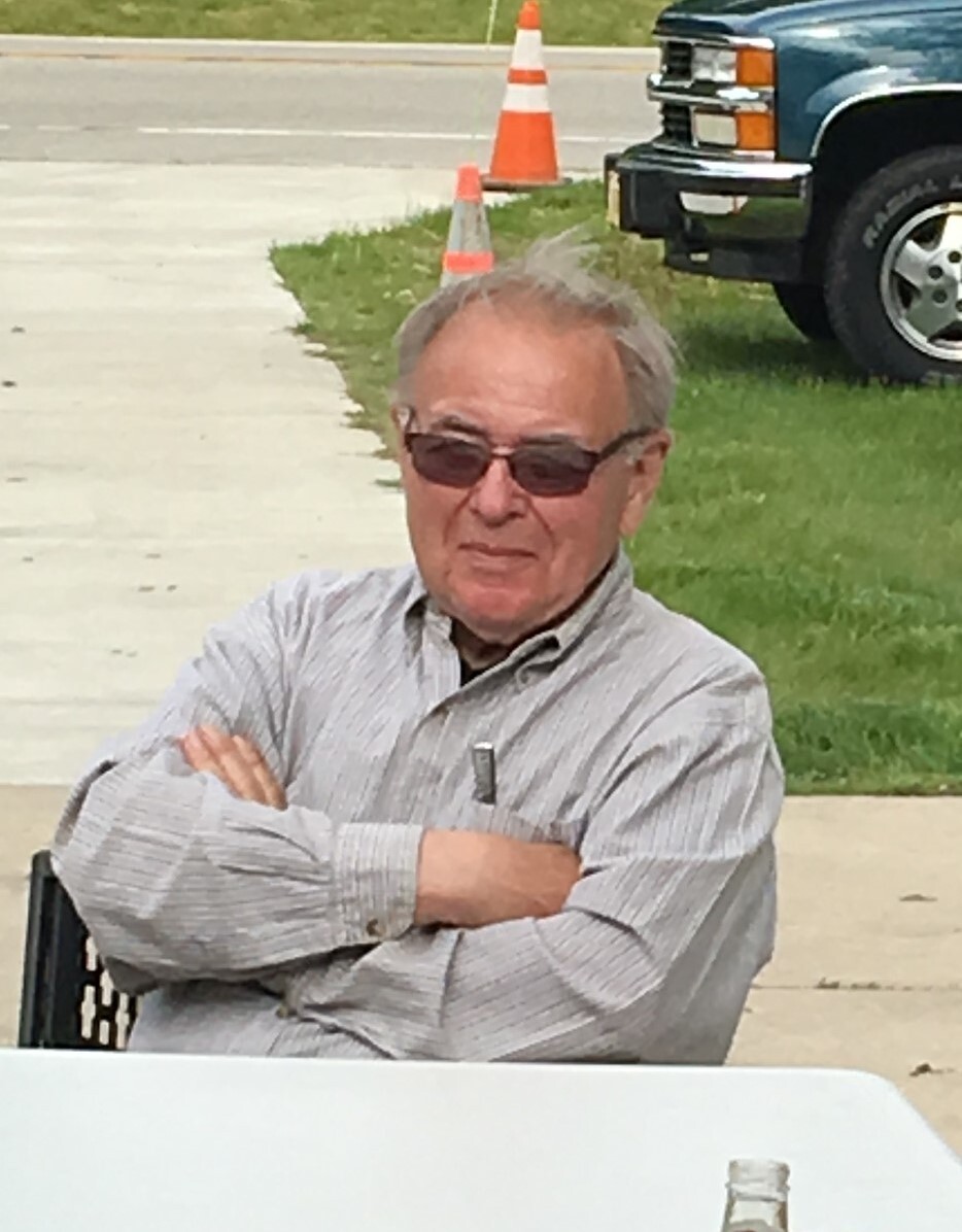 Older man with gray hair and sunglasses sits with arms crossed at a white table outdoors, traffic cones and a blue pickup truck in the background.