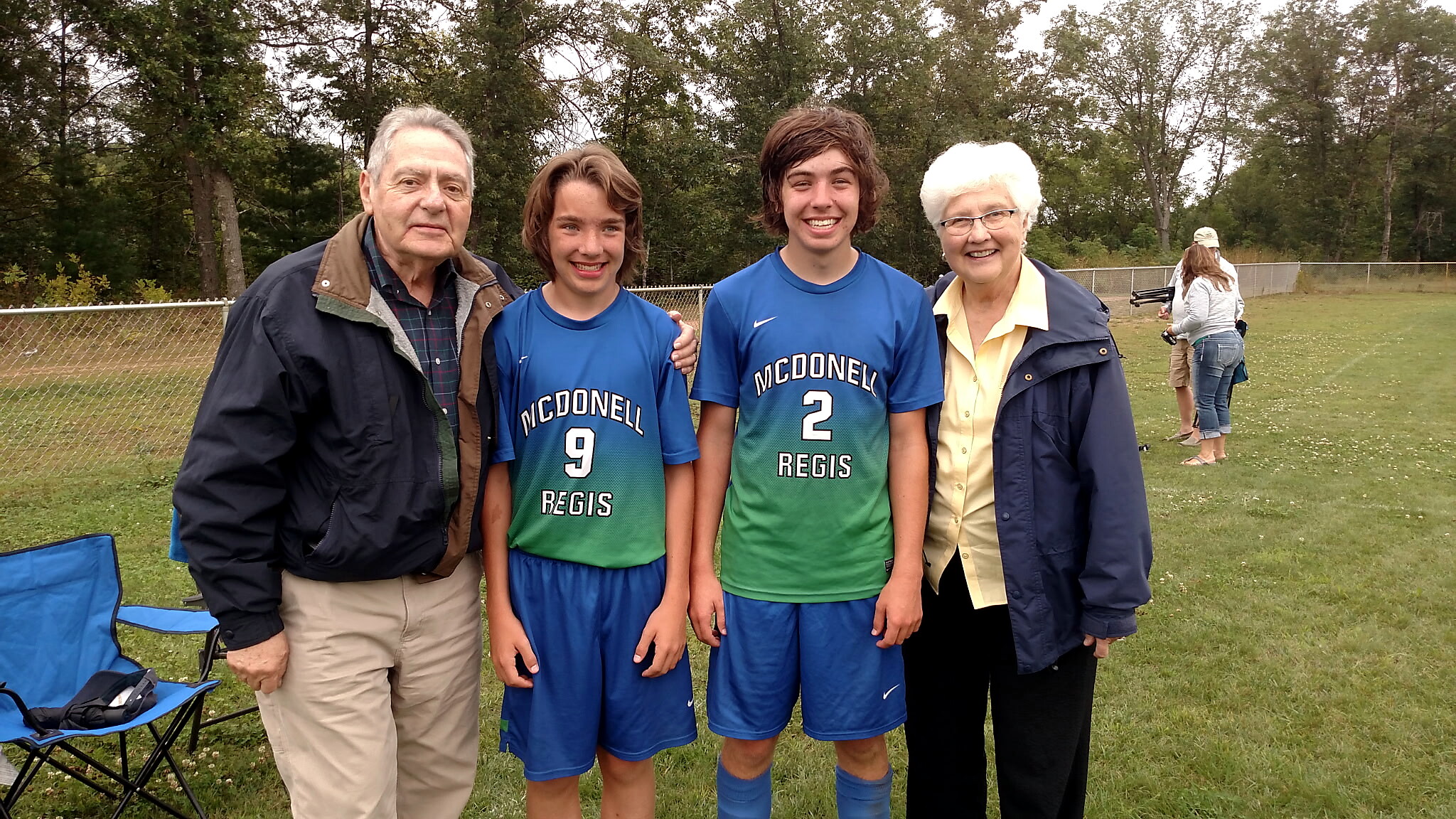 Group photo of two teenage boys in blue McDonell/Regis soccer uniforms with older adults on a grassy field, posing for the camera.