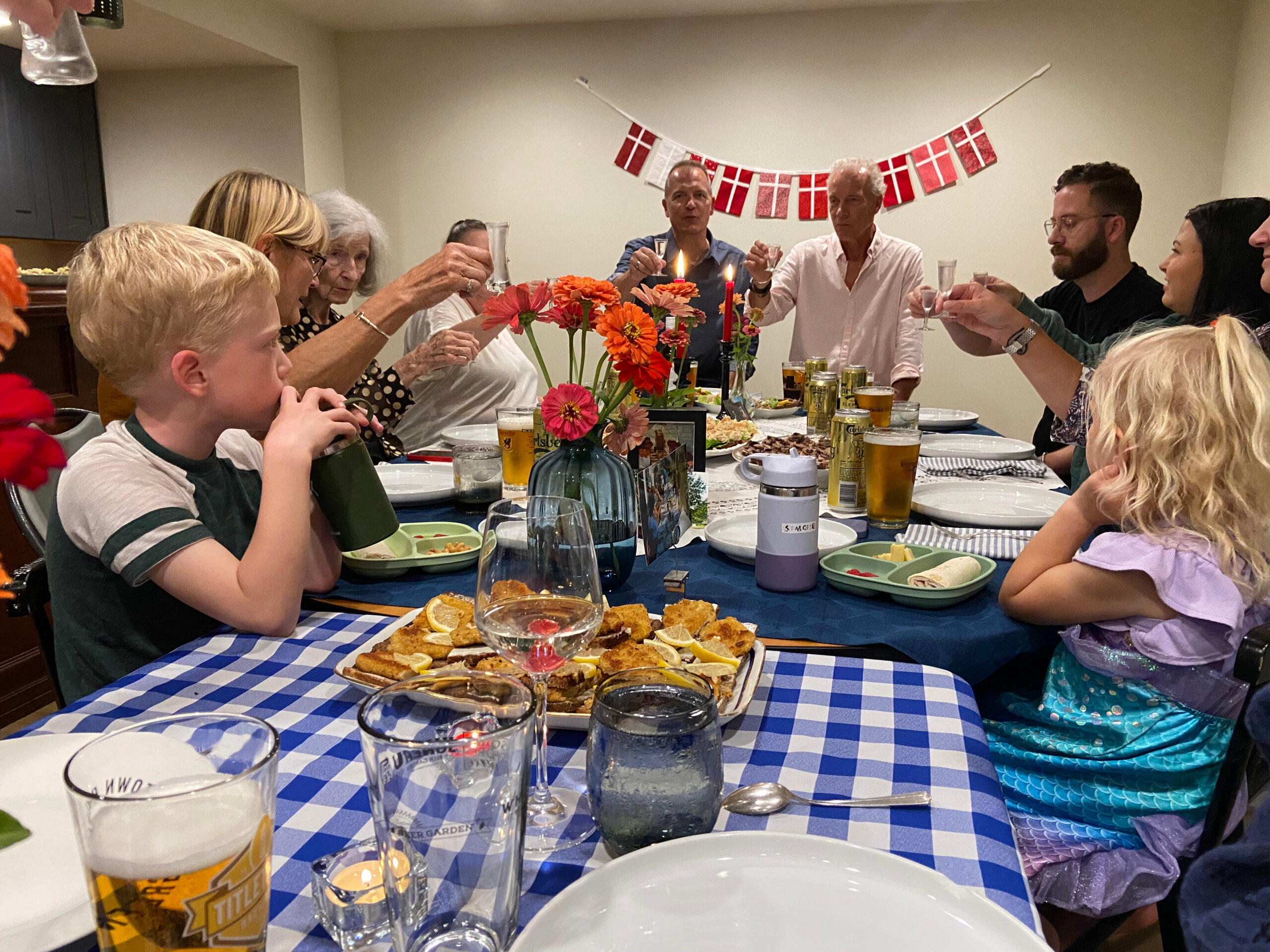 Group of people around a dining table toasting with glasses, Danish flag banner in background.