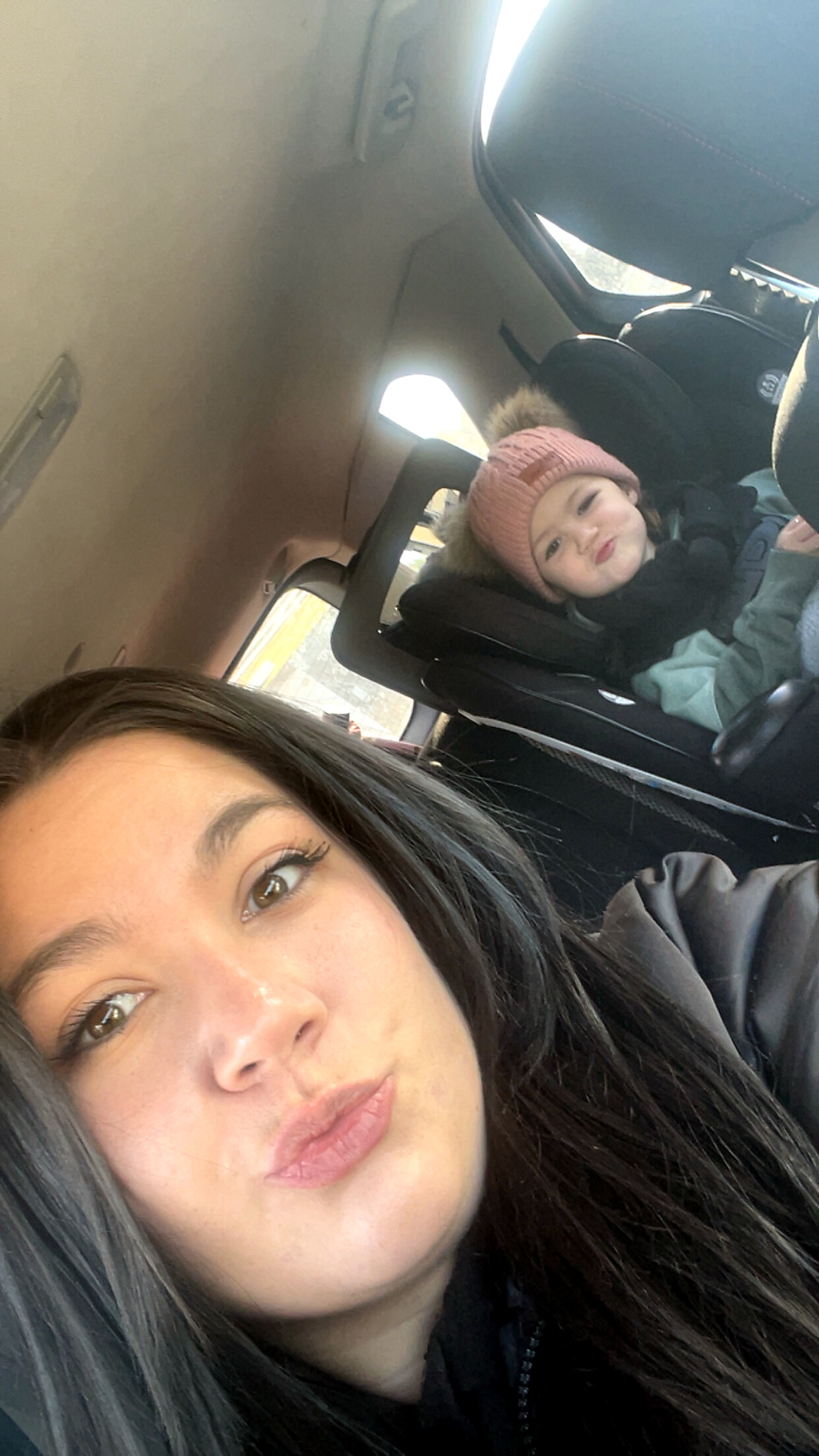 Woman in the foreground taking a selfie in a car, with a toddler in a car seat wearing a pink knit hat in the back seat.
