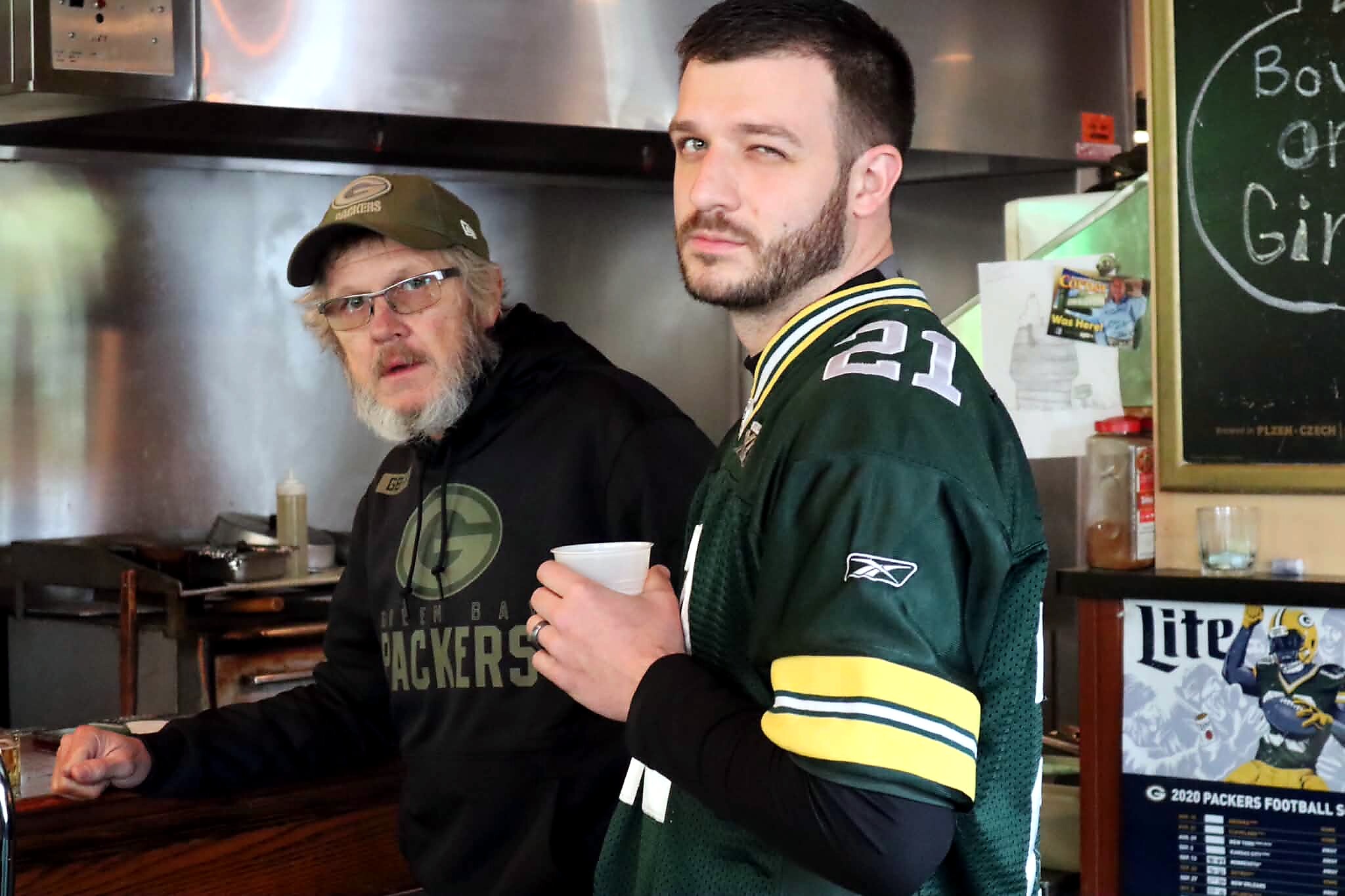 Two men in a kitchen or bar area, one in a Green Bay Packers jersey holding a cup and the other wearing a cap and glasses guarding the stove area behind them