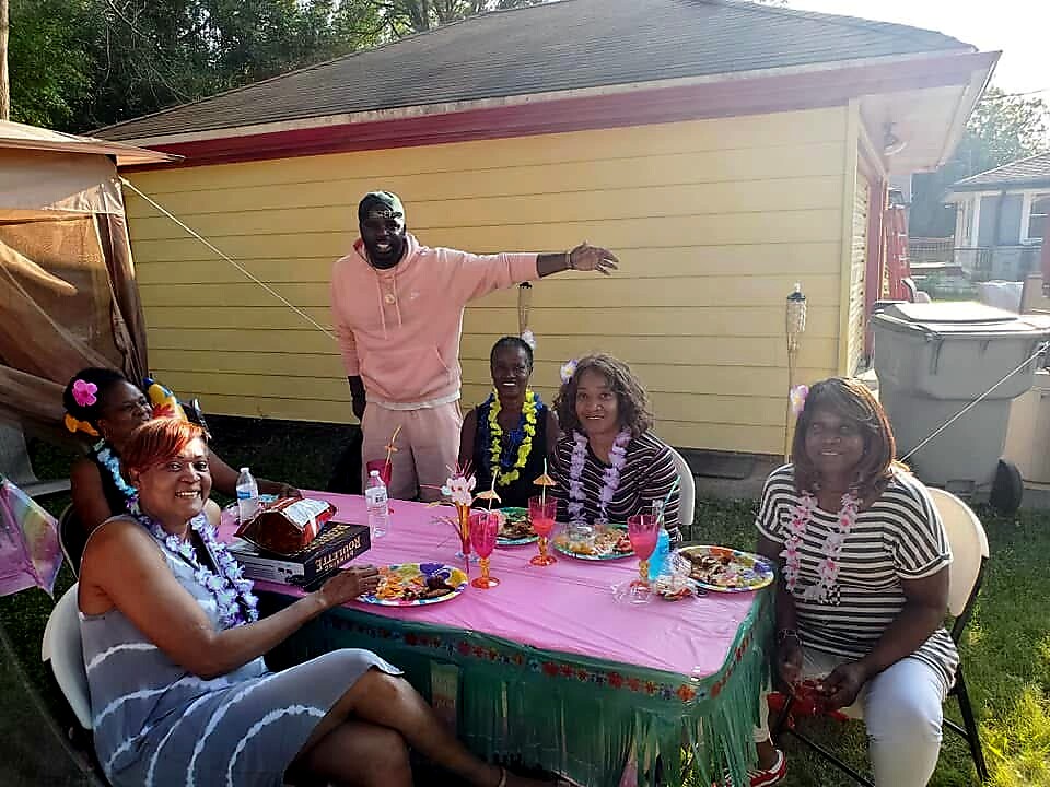 Five friends at a backyard birthday party around a pink table with leis and drinks; man in pink stands behind.