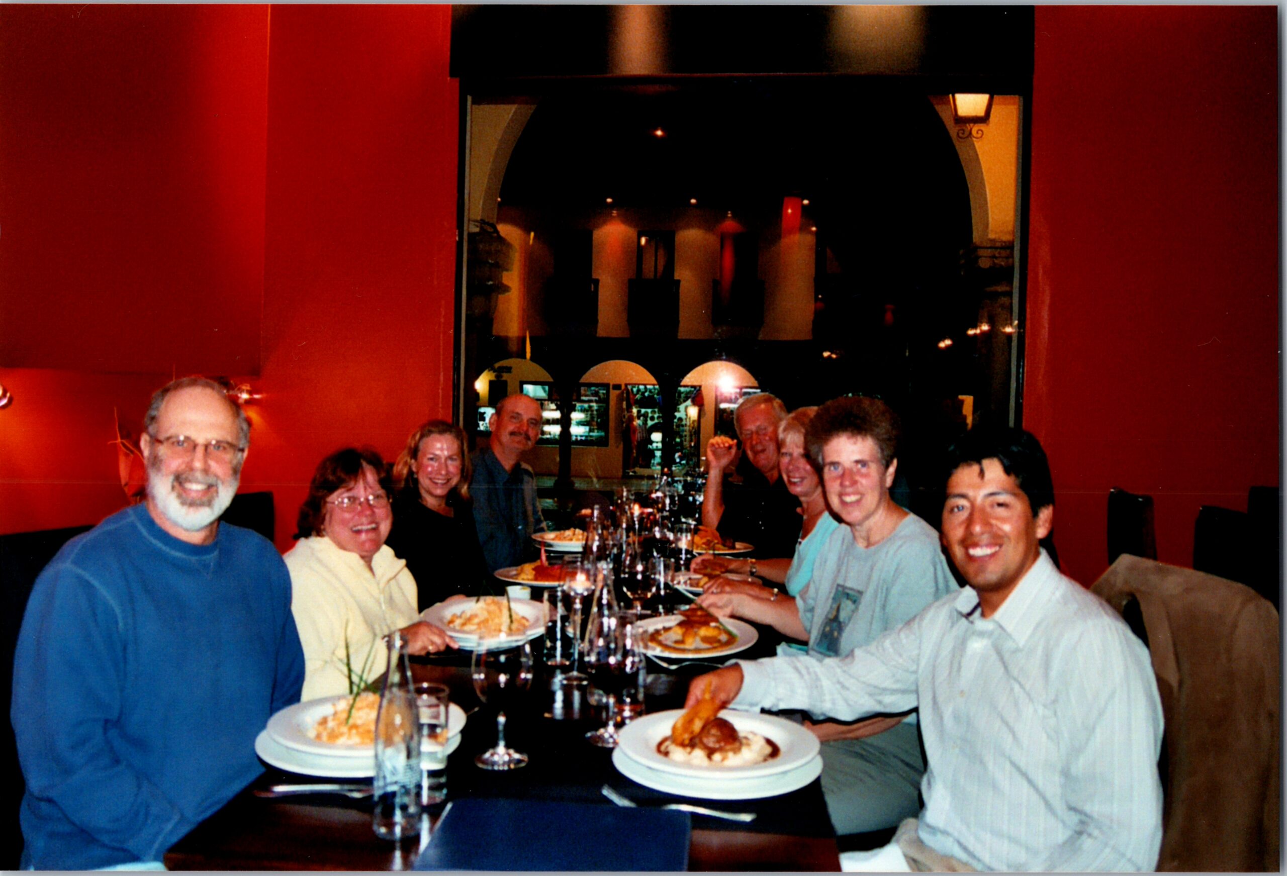 Nine people seated around a long table in a restaurant, plates of food and wine glasses in front of them, smiling at the camera as they share a meal.