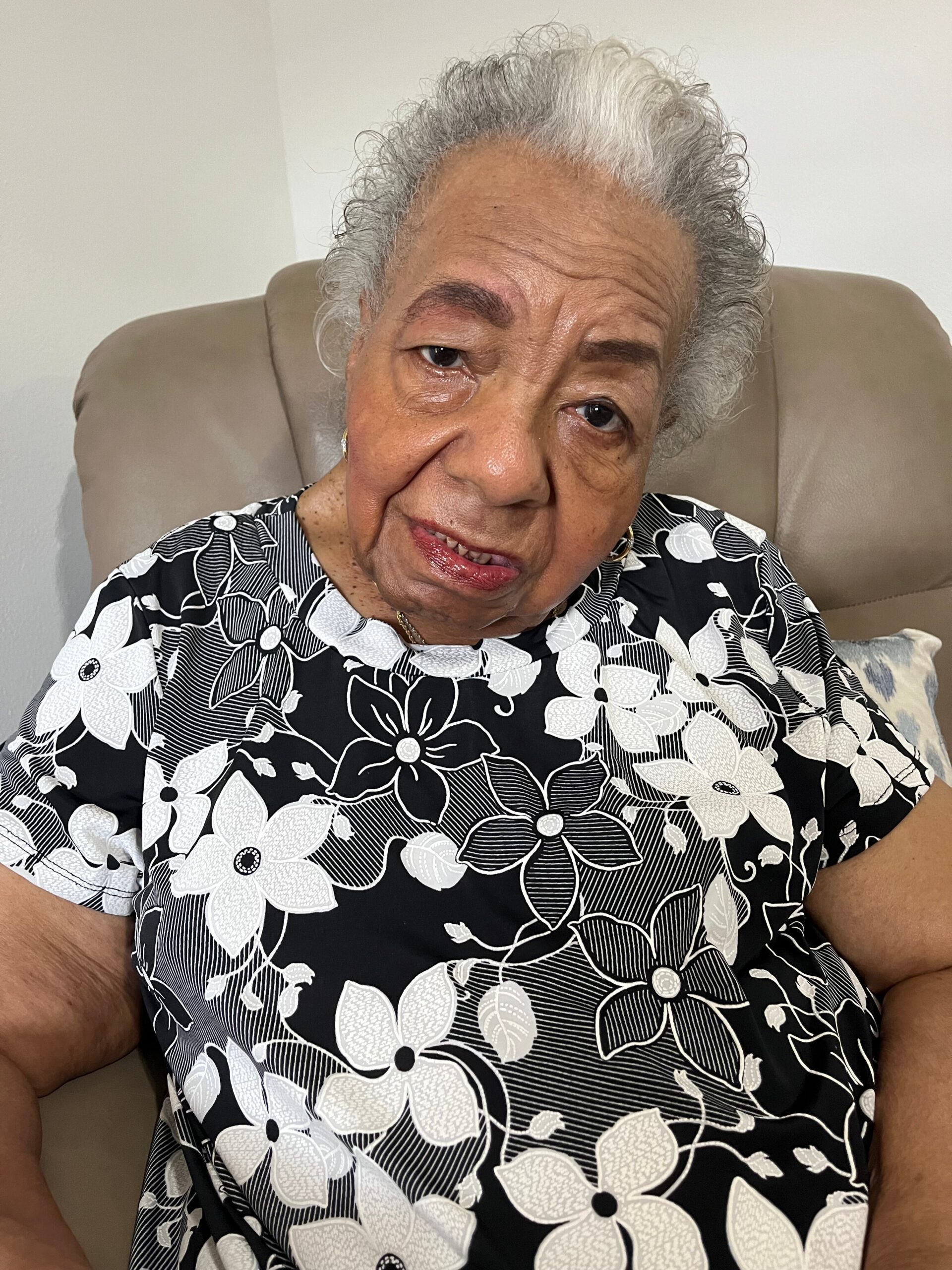 Close-up of an elderly woman with gray hair, seated on a beige chair, wearing a black-and-white floral blouse.